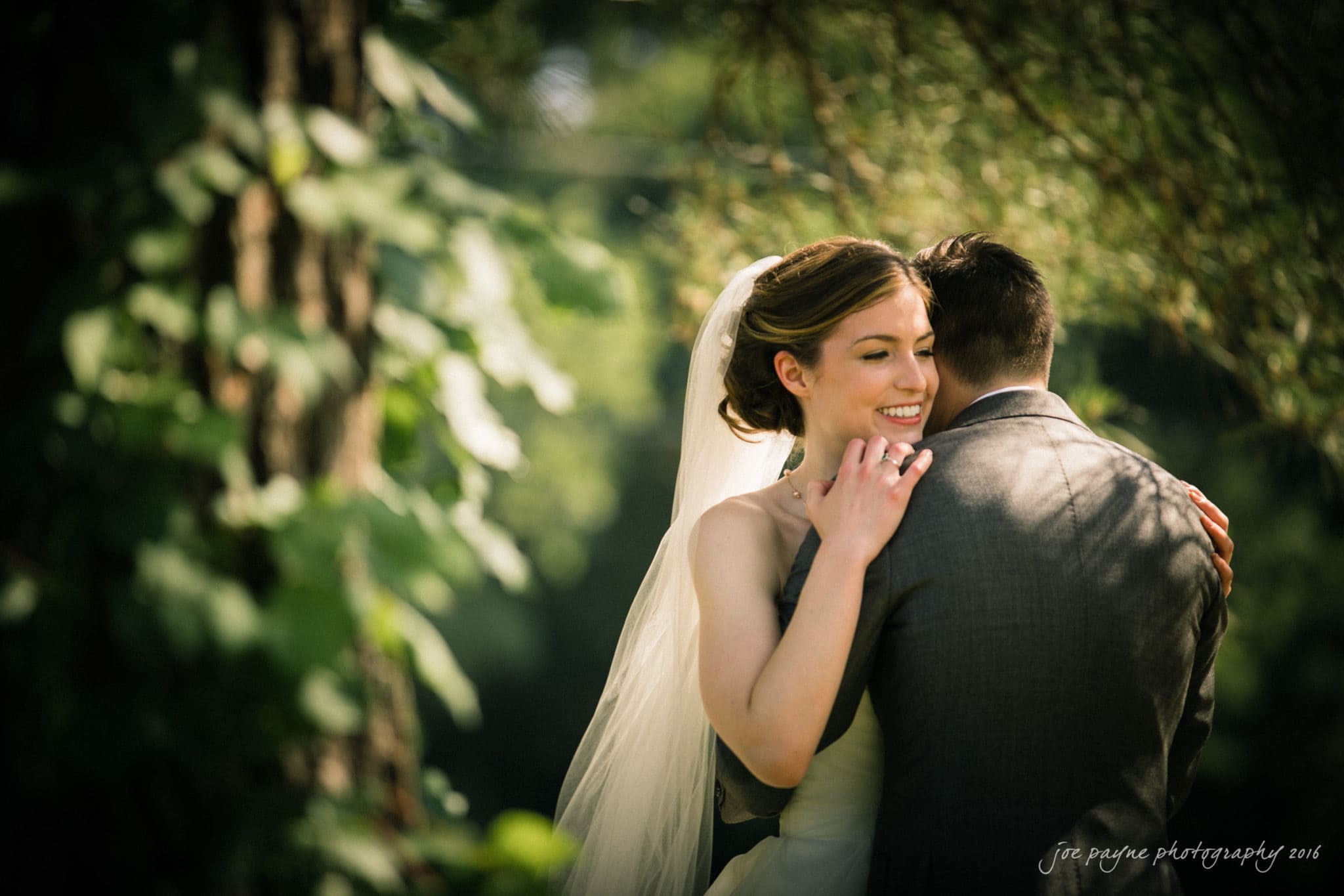 the oaks at salem wedding photography - kathryn & justin A bride in a white dress and veil embraces a groom in a gray suit outdoors. Sunlight filters through the trees, creating a warm, romantic atmosphere.