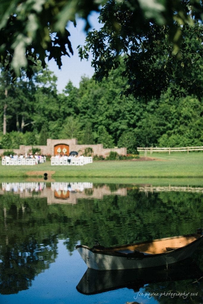 the oaks at salem wedding photography - kathryn & justin A rustic boat floats on a calm pond, reflecting the green trees and a distant wedding setup with white chairs and a floral backdrop. The scene is framed by leafy branches above.