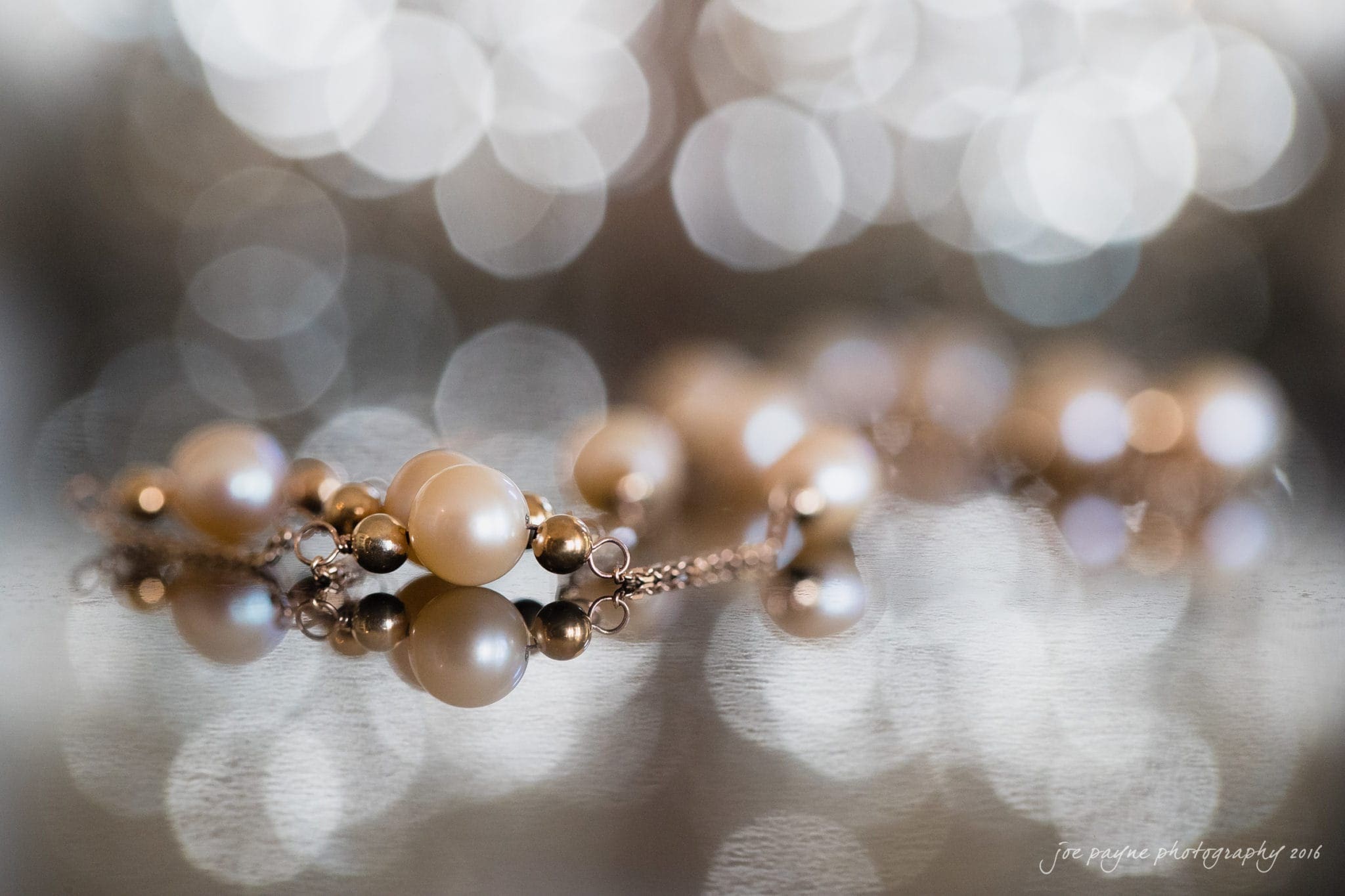 the oaks at salem wedding photography - kathryn & justin A close-up of a pearl necklace with gold beads, laid on a reflective surface. The background features soft, out-of-focus circular lights, creating a bokeh effect.