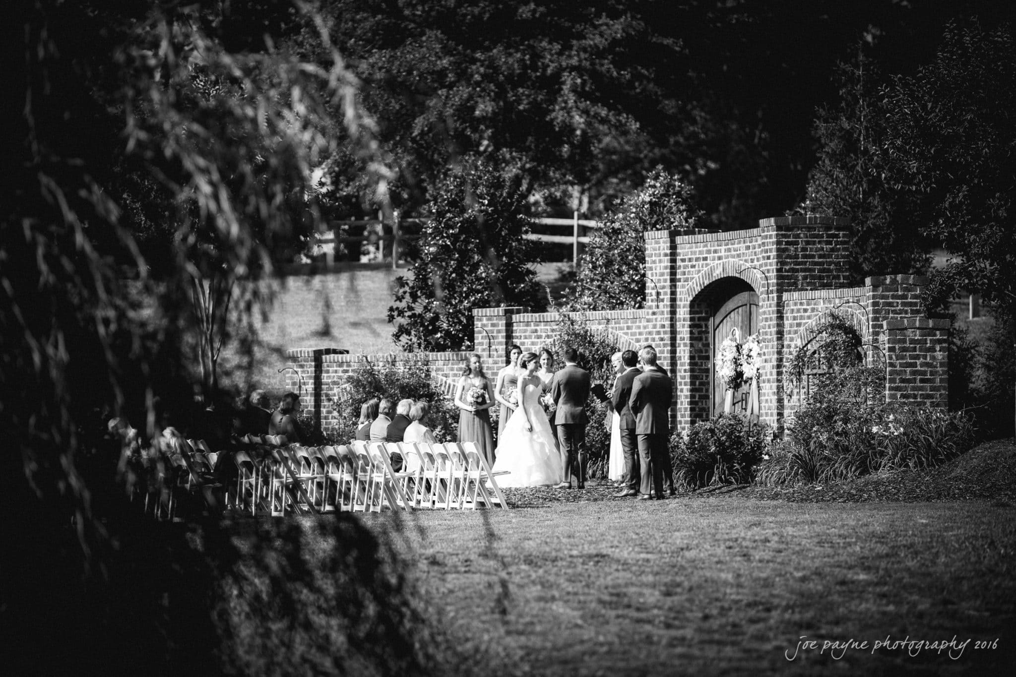 the oaks at salem wedding photography - kathryn & justin A black and white photo of an outdoor wedding ceremony, featuring a bride and groom standing with an officiant by a brick archway. Guests are seated in rows on the left, surrounded by lush greenery.