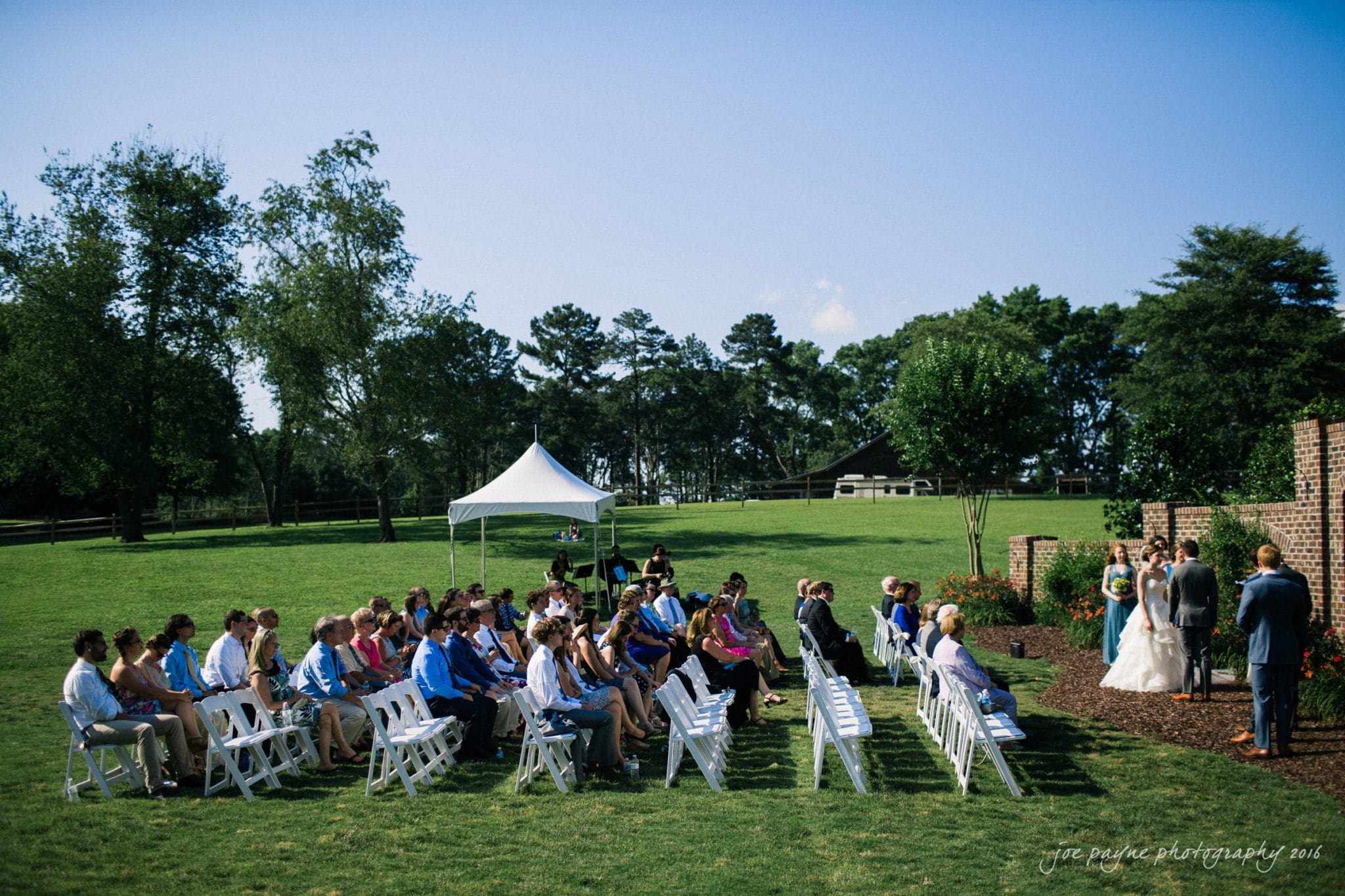 the oaks at salem wedding photography - kathryn & justin A wedding ceremony in an outdoor garden setting, with guests seated on white chairs. The bride and groom stand together near a brick structure, with trees and a white tent visible in the grassy background. Its a sunny day.