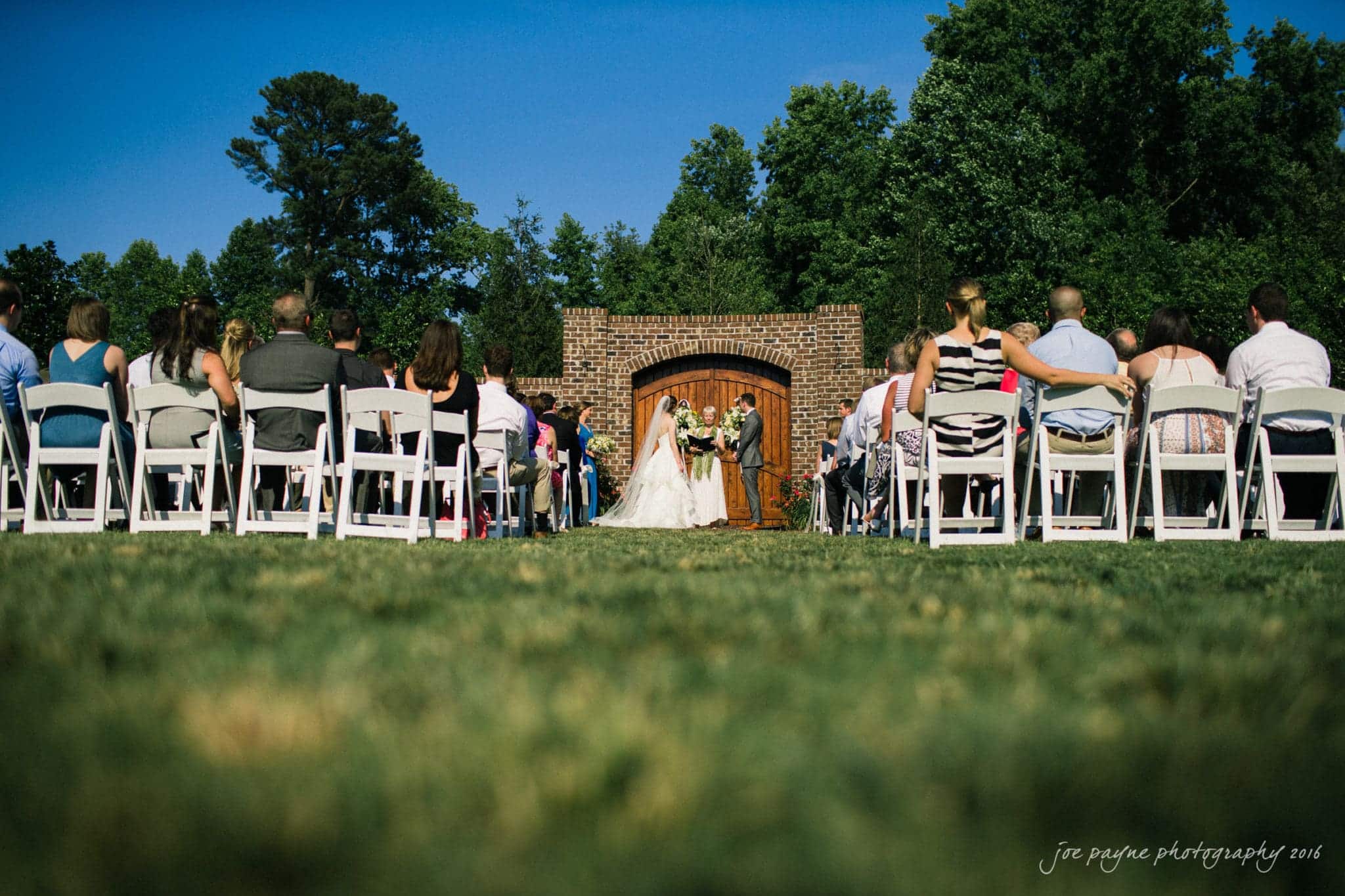 the oaks at salem wedding photography - kathryn & justin A bride and groom stand at an outdoor altar, framed by a brick archway, during a wedding ceremony. Guests are seated on white chairs, facing the couple. The setting is lush with green grass and trees under a clear blue sky.