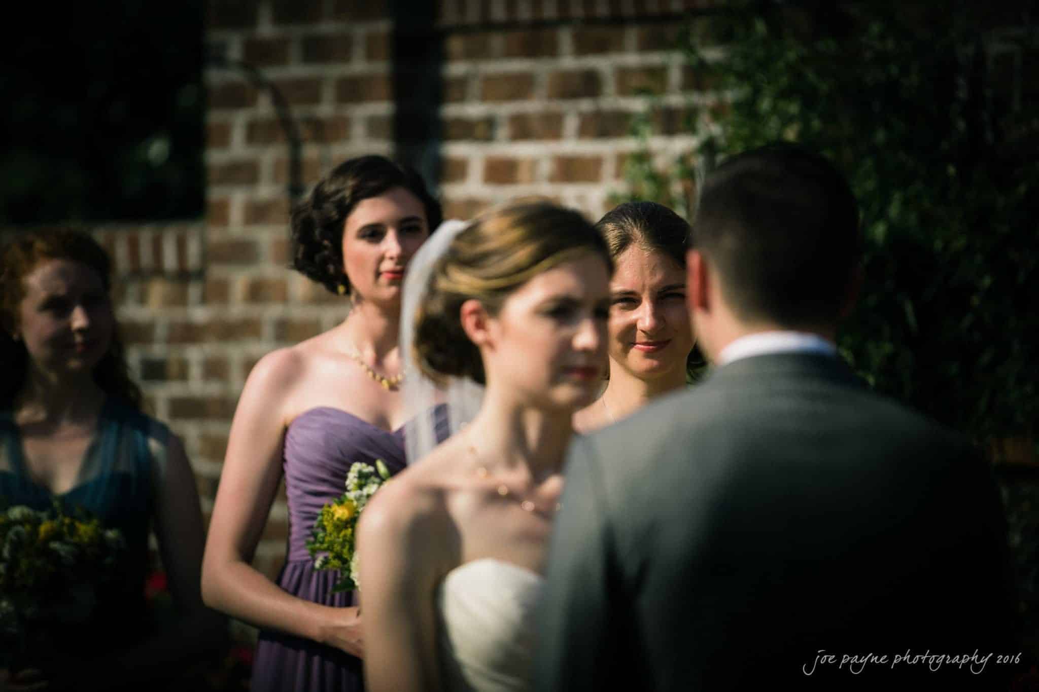the oaks at salem wedding photography - kathryn & justin A bride and groom stand facing each other during an outdoor wedding ceremony. Bridesmaids in colorful dresses are in the background, with one holding flowers. They are all standing in front of a brick wall.
