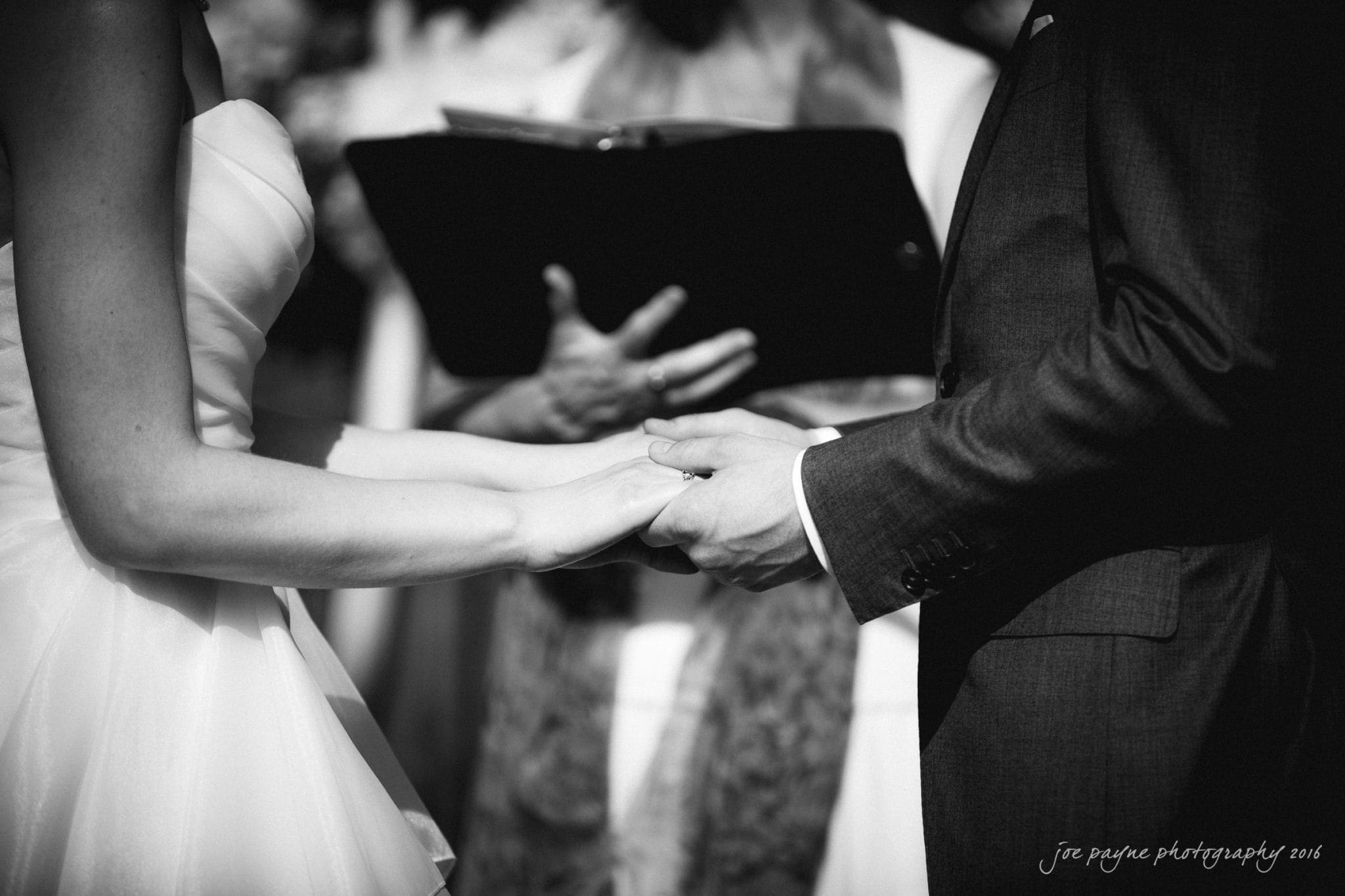 the oaks at salem wedding photography - kathryn & justin A bride and groom hold hands during a wedding ceremony. They stand facing each other in formal attire, with the officiant holding a book in the background. The image has a soft focus, emphasizing the emotion of the moment.