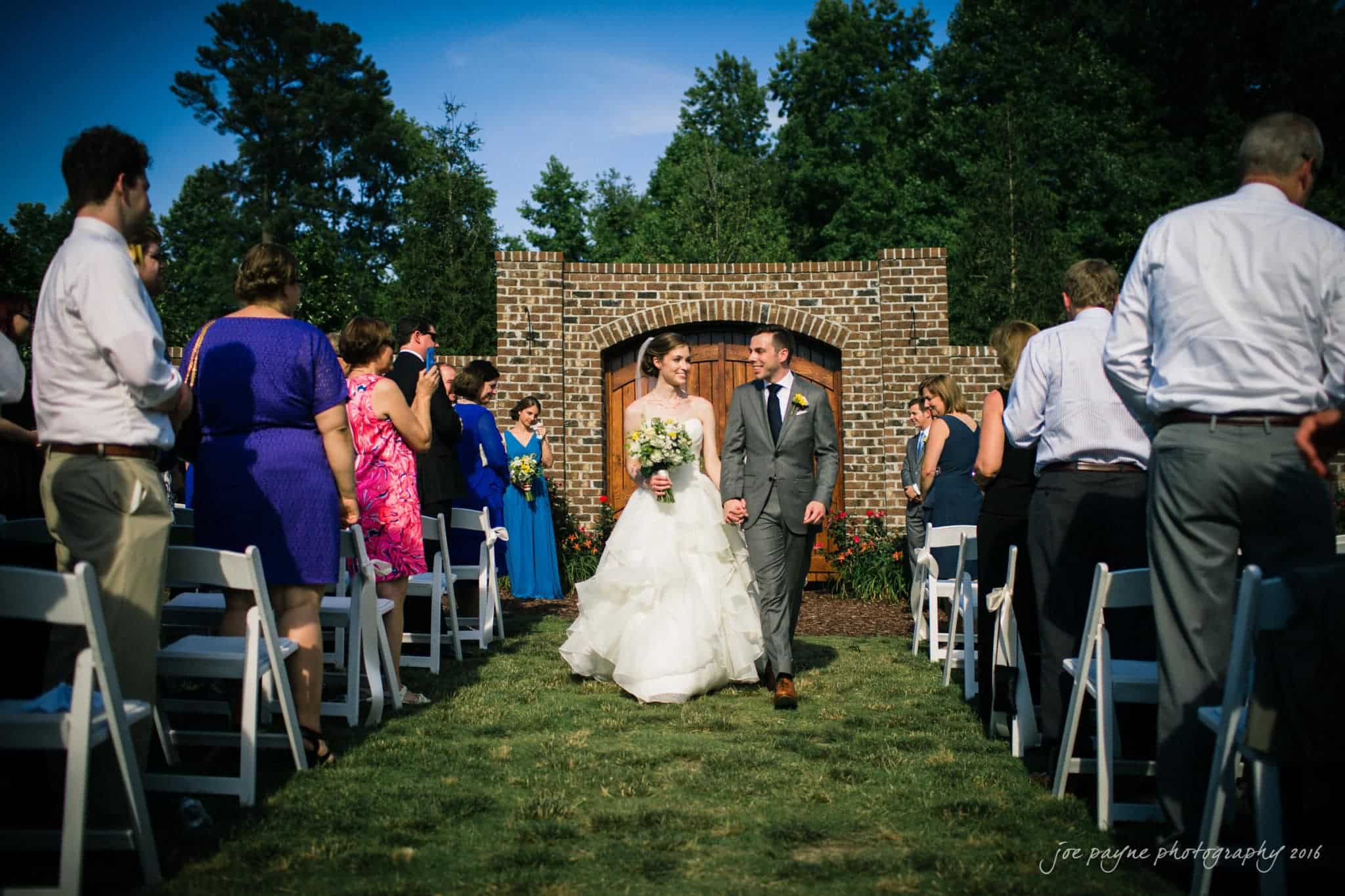 the oaks at salem wedding photography - kathryn & justin A bride and groom walk down the aisle together outdoors, smiling and holding hands. Guests stand and applaud on either side. The background features a brick structure and trees under a clear blue sky.