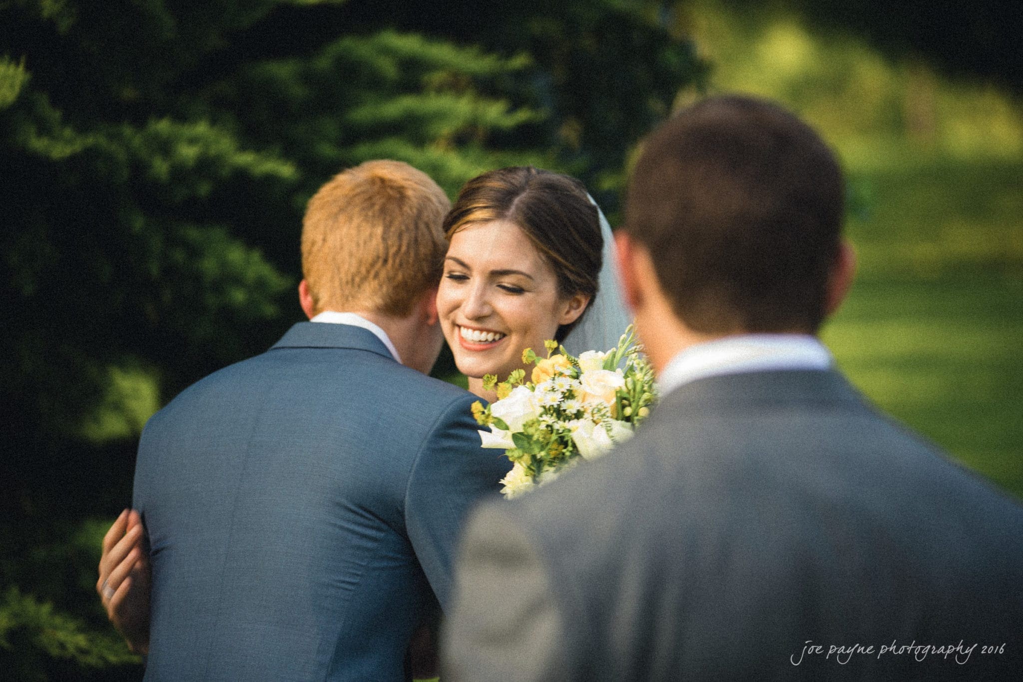 the oaks at salem wedding photography - kathryn & justin A bride holding a bouquet smiles while hugging a man in a blue suit at an outdoor event, with another man standing in the foreground. Lush greenery is visible in the background.