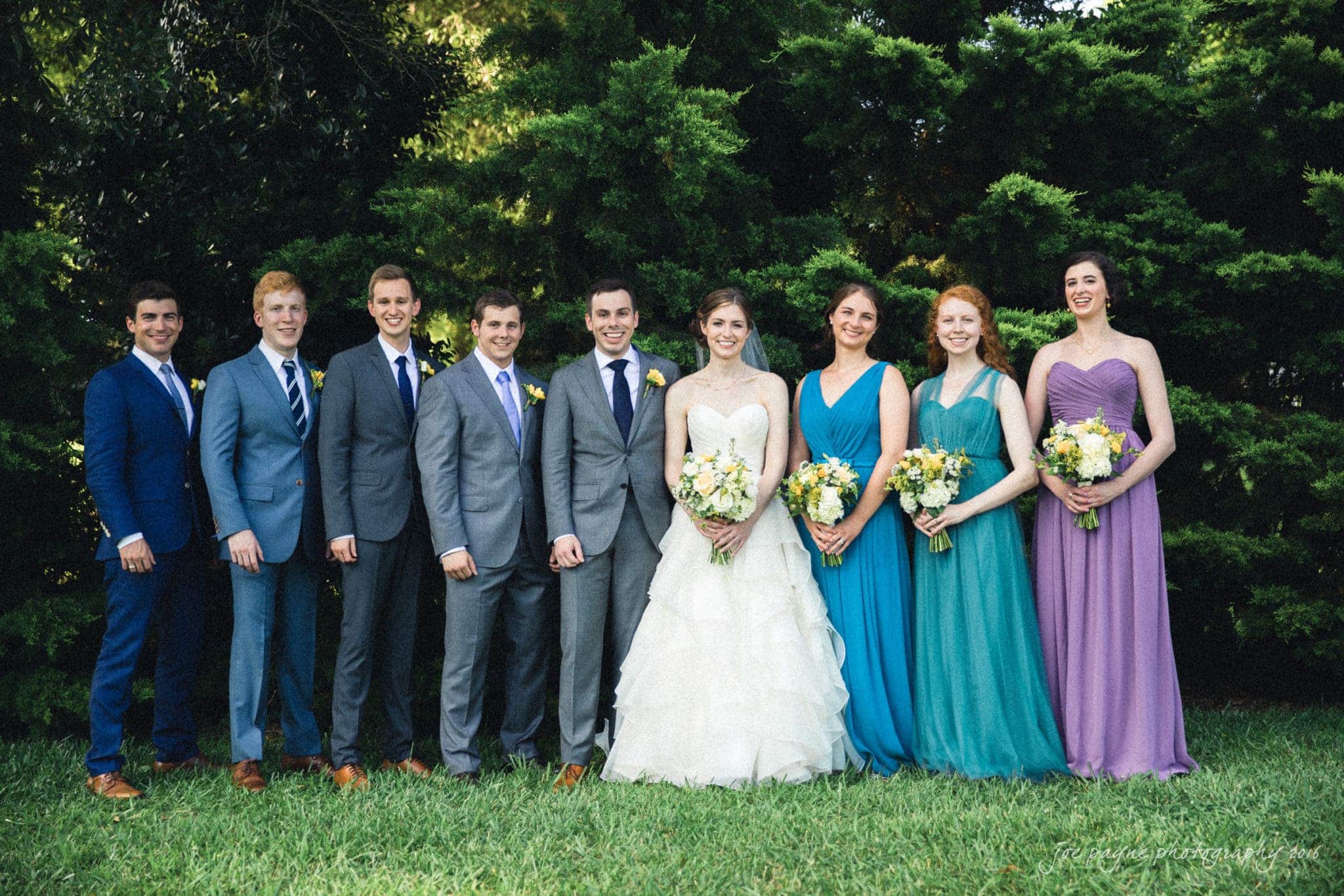 the oaks at salem wedding photography - kathryn & justin A wedding party with nine people standing in a row. The bride is in a white gown, holding a bouquet. The groom and groomsmen wear suits in various colors. The bridesmaids wear dresses in shades of blue and purple, each holding a bouquet. Trees are in the background.