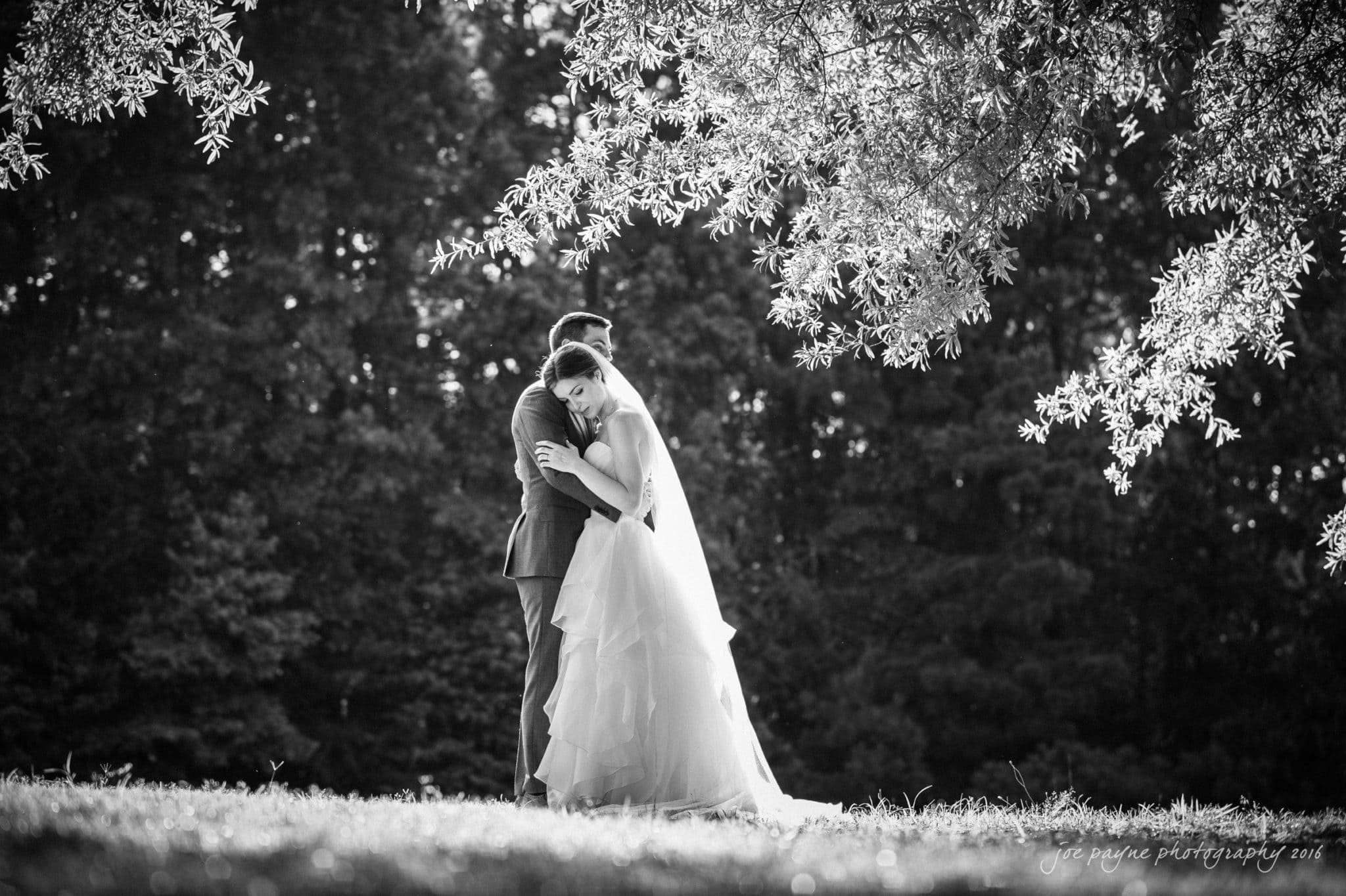 the oaks at salem wedding photography - kathryn & justin A black and white photo of a bride and groom embracing under a tree. The bride is wearing a long, flowing dress and veil, and the groom is in a suit. Sunlight filters through the leaves, creating a serene and romantic atmosphere.