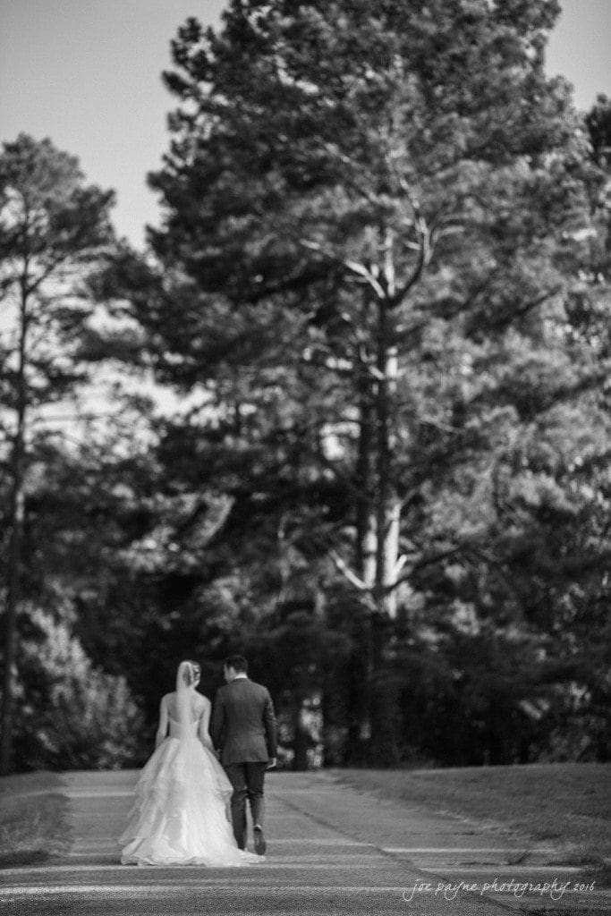 the oaks at salem wedding photography - kathryn & justin A bride and groom walk hand in hand down a paved path, framed by tall trees. The bride is in a flowing gown, and the groom is in a suit. The setting is serene and natural, captured in black and white.