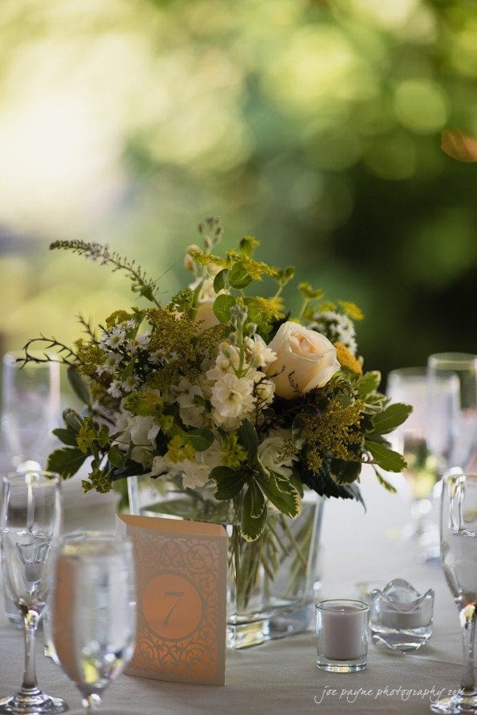 the oaks at salem wedding photography - kathryn & justin A table centerpiece with a bouquet of white and yellow flowers in a square glass vase. A small card with the number 7 is placed next to it. Glasses and tealight candles surround the arrangement on a white tablecloth.