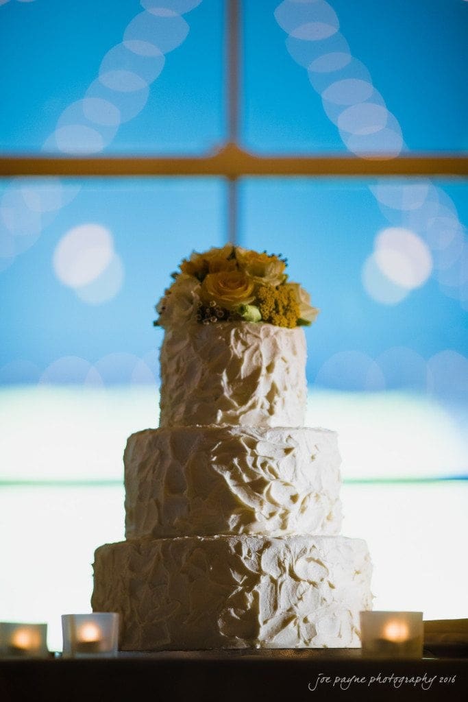 the oaks at salem wedding photography - kathryn & justin A three-tiered white wedding cake with textured frosting and a bouquet of yellow roses on top. The background is softly blurred, with twinkling lights and a vivid blue hue, creating a romantic ambiance. Three small candles surround the cake.