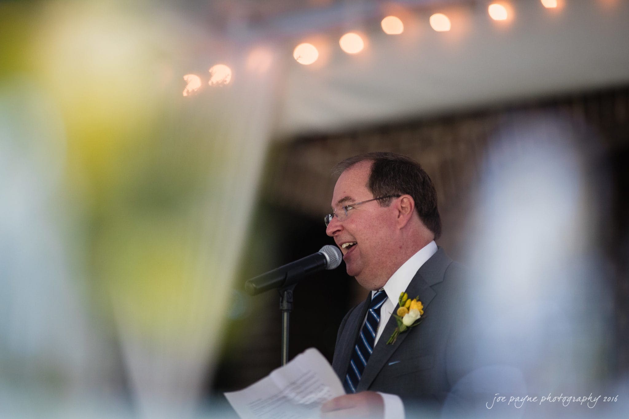 the oaks at salem wedding photography - kathryn & justin A man in a suit and striped tie speaks into a microphone, smiling as he holds a piece of paper. A yellow flower is pinned to his lapel. The background is softly lit with string lights.