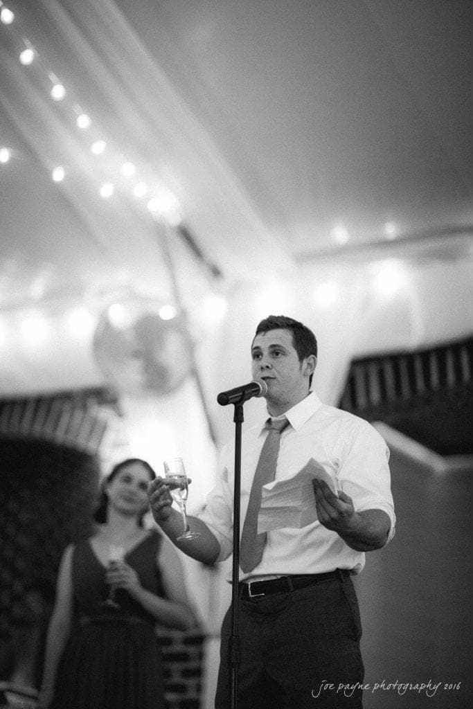 the oaks at salem wedding photography - kathryn & justin A man in a white shirt and tie is speaking into a microphone, holding a piece of paper and a glass. A woman in a dress stands behind him. The setting appears to be a formal event with string lights and draped fabric in the background.