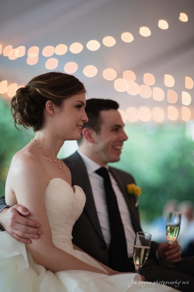 the oaks at salem wedding photography - kathryn & justin A bride and groom are sitting close together, smiling and holding glasses of champagne. They are under soft, warm string lights, creating a romantic atmosphere. The groom has his arm around the bride.