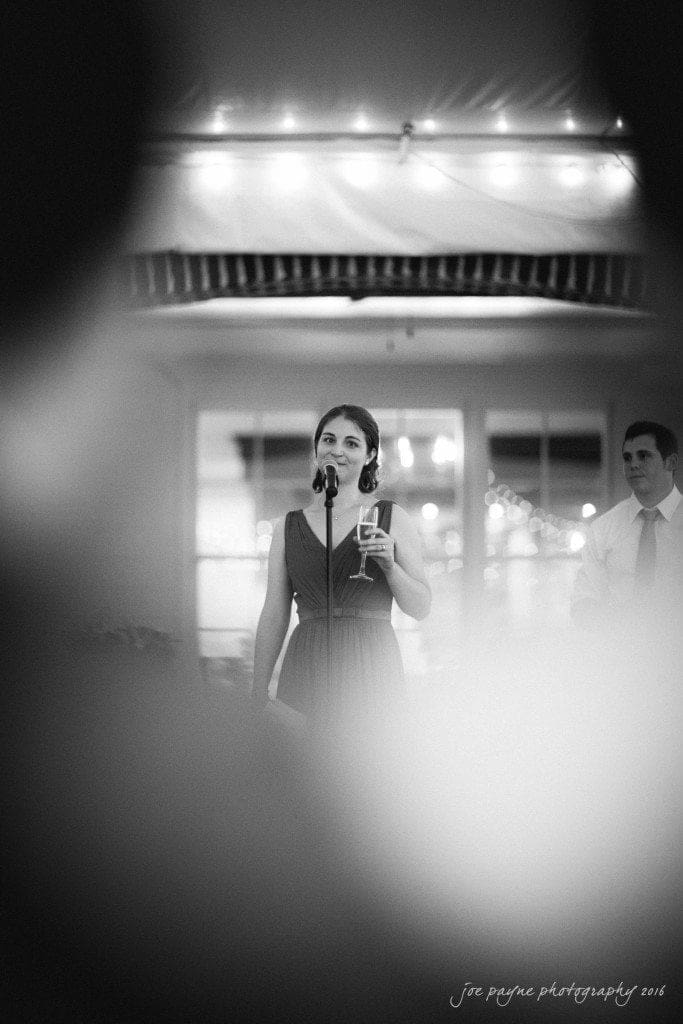 the oaks at salem wedding photography - kathryn & justin A woman in a dress holds a glass and speaks into a microphone, smiling warmly. Soft lights create a gentle, festive ambiance. A man in a tie stands in the background, partially visible. The image has a black and white filter.