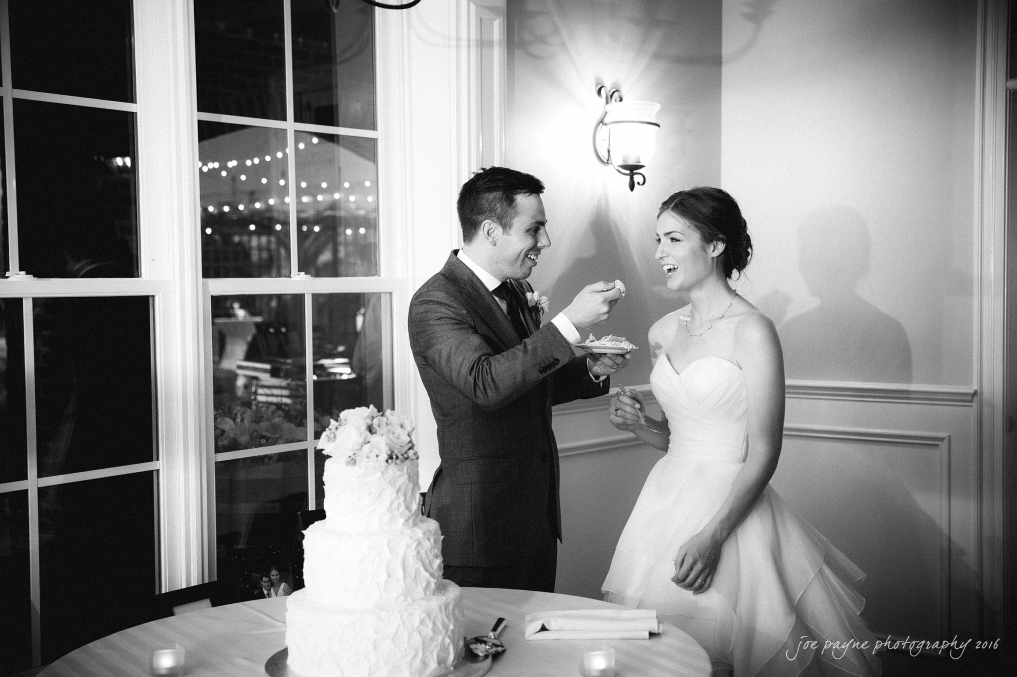 the oaks at salem wedding photography - kathryn & justin A groom in a suit feeds a smiling bride in a strapless dress a piece of wedding cake. They stand beside a table with a large tiered cake. The room is softly lit, and there are fairy lights visible through a window.