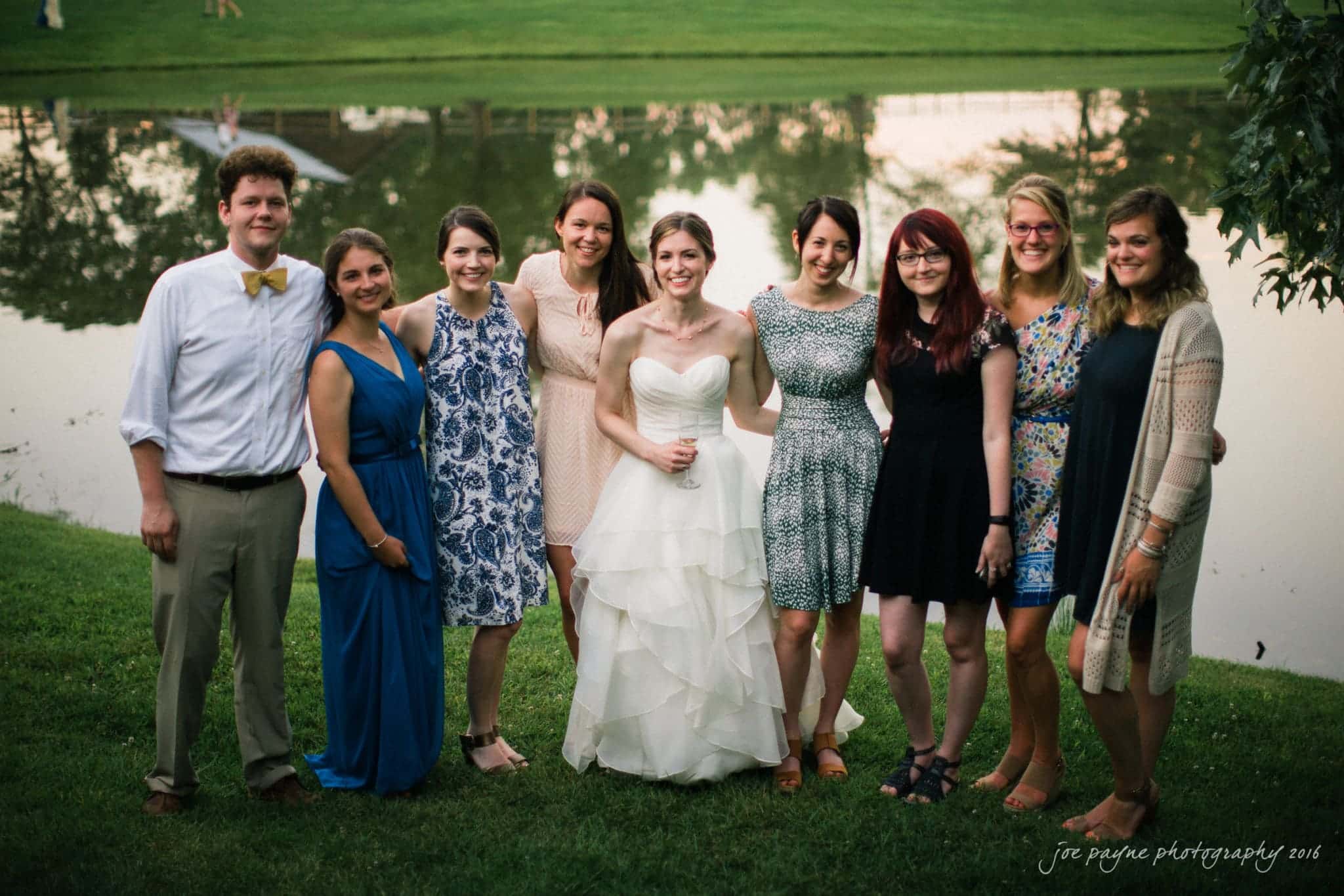 the oaks at salem wedding photography - kathryn & justin A bride in a white dress stands with a group of friends in front of a pond. The women wear various dresses and one man wears a shirt and tie. They are smiling and standing on grass with trees reflected in the pond behind them.