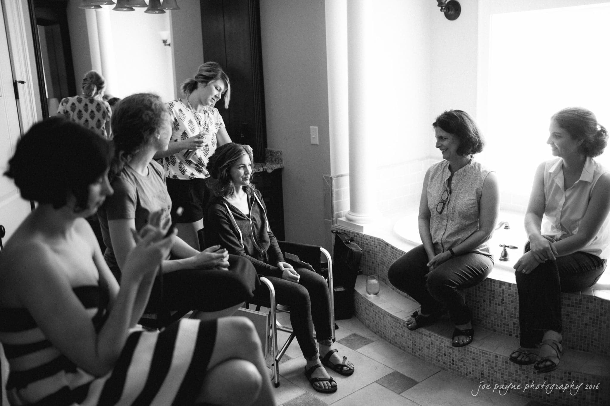 the oaks at salem wedding photography - kathryn & justin A group of women are gathered in a room with large windows. One woman sits in a chair having her hair styled, while others sit and stand around casually, talking and using their phones. The room appears relaxed and well-lit.