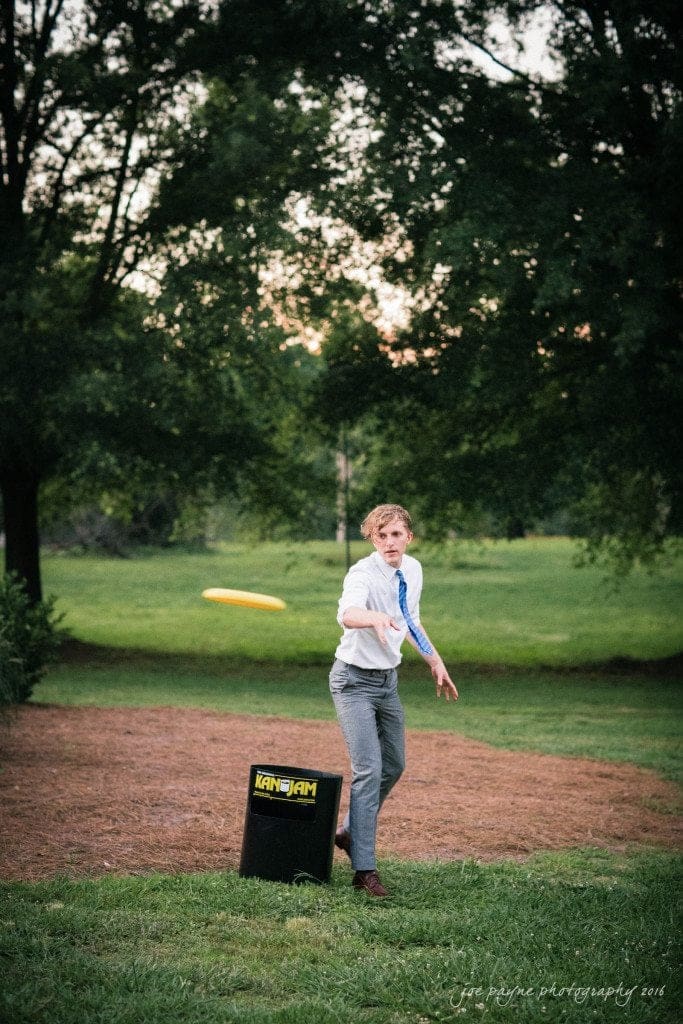 the oaks at salem wedding photography - kathryn & justin A person in a tie and rolled-up sleeves is playing KanJam outdoors, aiming a frisbee toward a black KanJam goal. The setting is a grassy area with trees in the background.