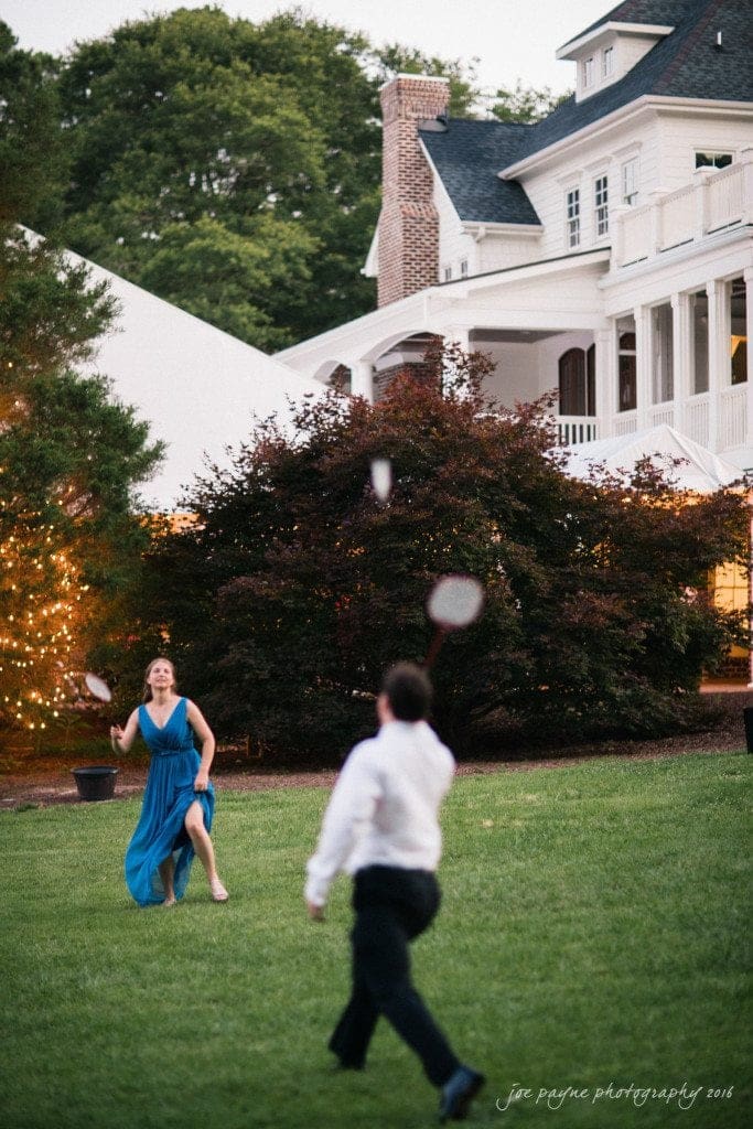 the oaks at salem wedding photography - kathryn & justin Two people play badminton on a grassy lawn in front of a large house. The woman in a blue dress is in the foreground, looking focused on the game. The house is white with multiple windows and a brick chimney. Trees surround the area.