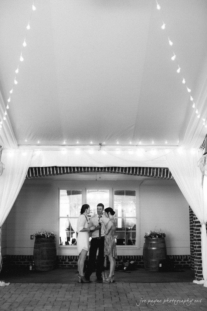 the oaks at salem wedding photography - kathryn & justin A black and white photo shows three people standing under a tent with soft string lights. They are having a conversation and holding drinks. Two wooden barrels are visible in the background against a brick wall.