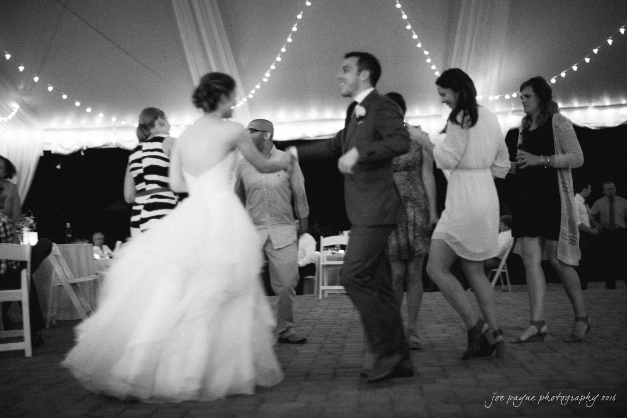 the oaks at salem wedding photography - kathryn & justin A black and white photo of people dancing joyfully at an outdoor event under a tent with string lights. A person in a gown and a person in a suit are prominent in the foreground, with other guests chatting and enjoying the scene.