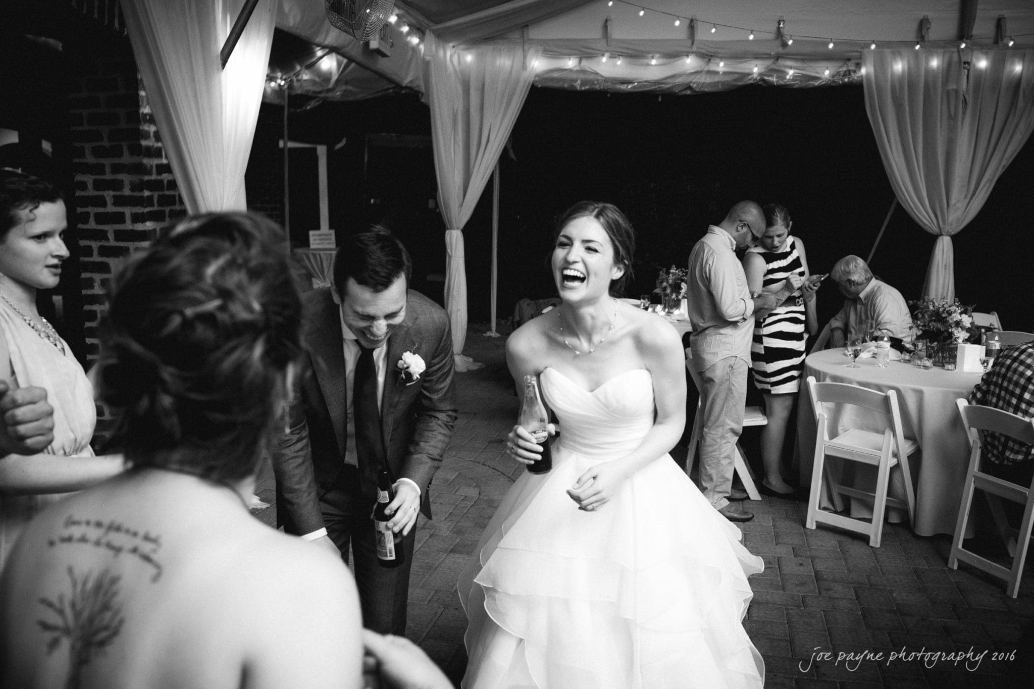 the oaks at salem wedding photography - kathryn & justin A joyful bride in a strapless gown laughs with guests at an outdoor wedding reception. People are gathered under a tent with string lights, sitting at tables and socializing. Black and white photo.