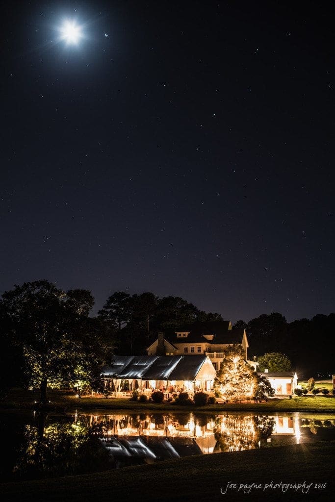 the oaks at salem wedding photography - kathryn & justin A serene nighttime scene with a bright moon and starry sky above a well-lit house by a reflective pond. Trees surround the house, and the calm water mirrors the tranquil atmosphere.