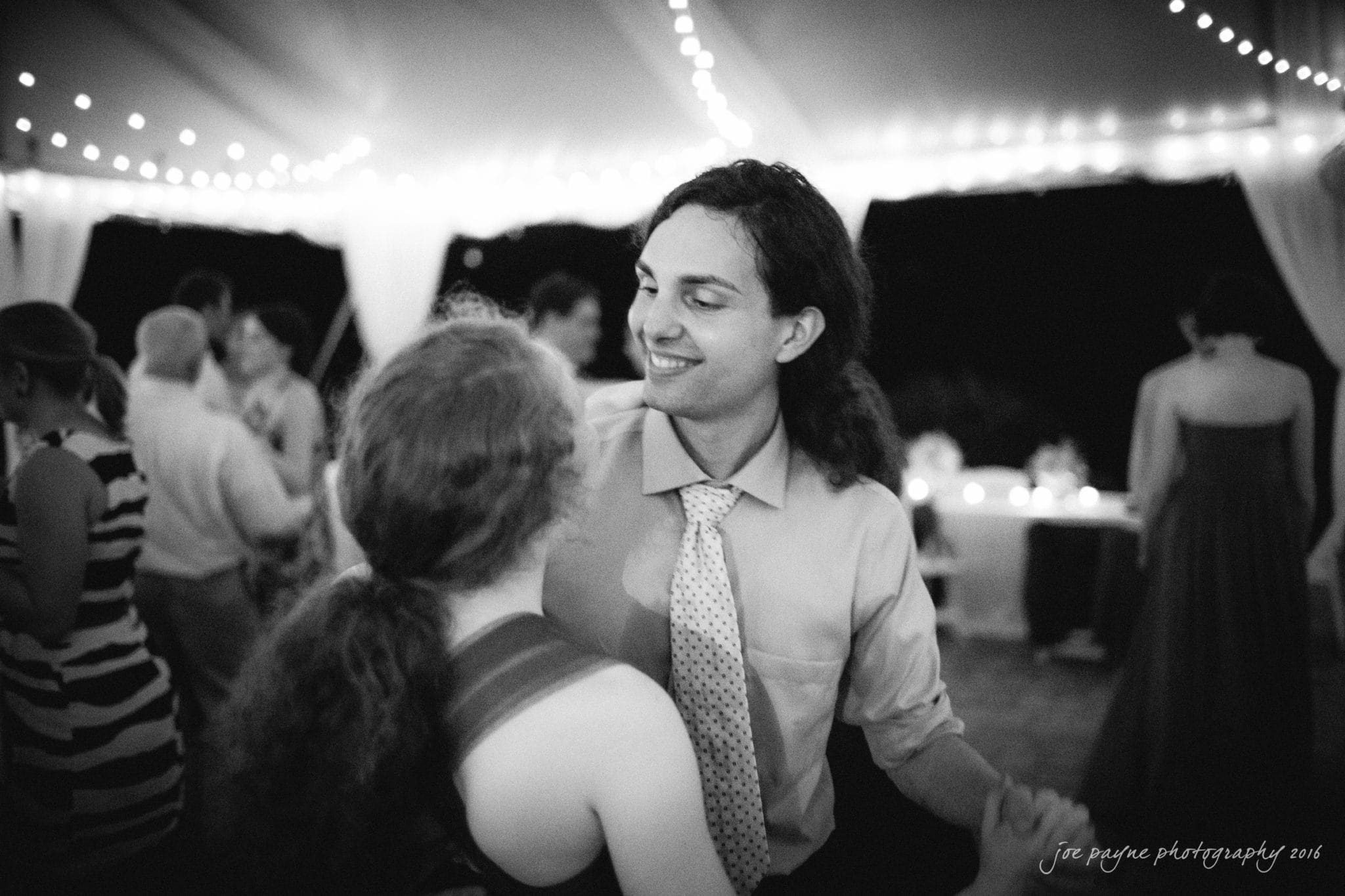 the oaks at salem wedding photography - kathryn & justin A couple dances under a canopy with string lights at an outdoor evening event. The man, with long hair and wearing a tie, smiles at his partner, who has her hair tied back. Other people are seen dancing and mingling in the background. Black and white photo.