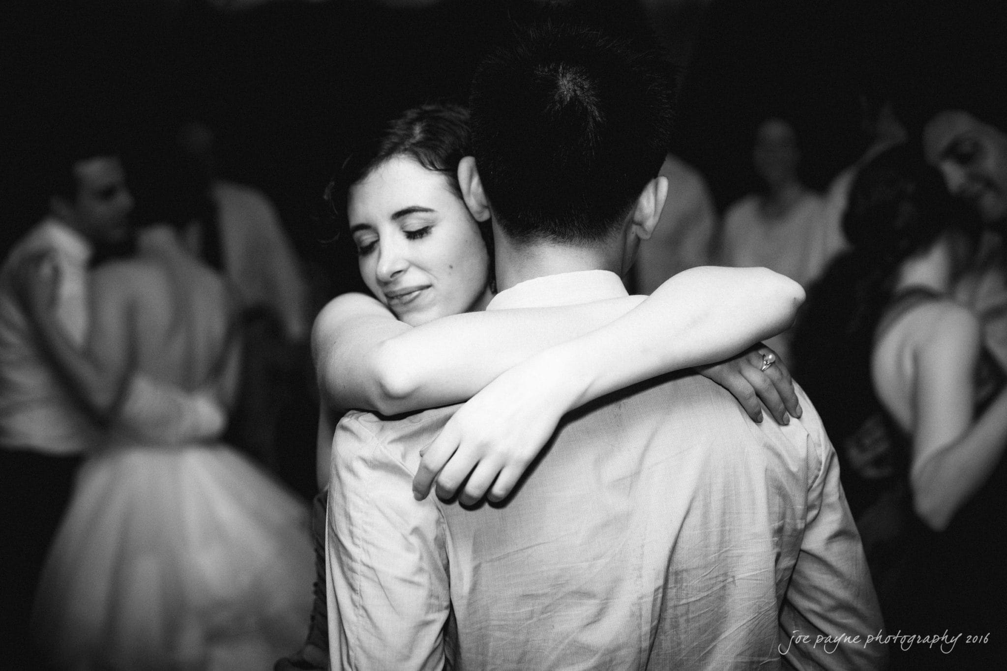 the oaks at salem wedding photography - kathryn & justin A black and white photo of a couple embracing on a dance floor. The woman has her eyes closed and smiles softly, while holding the man. Other people are visible dancing in the background.