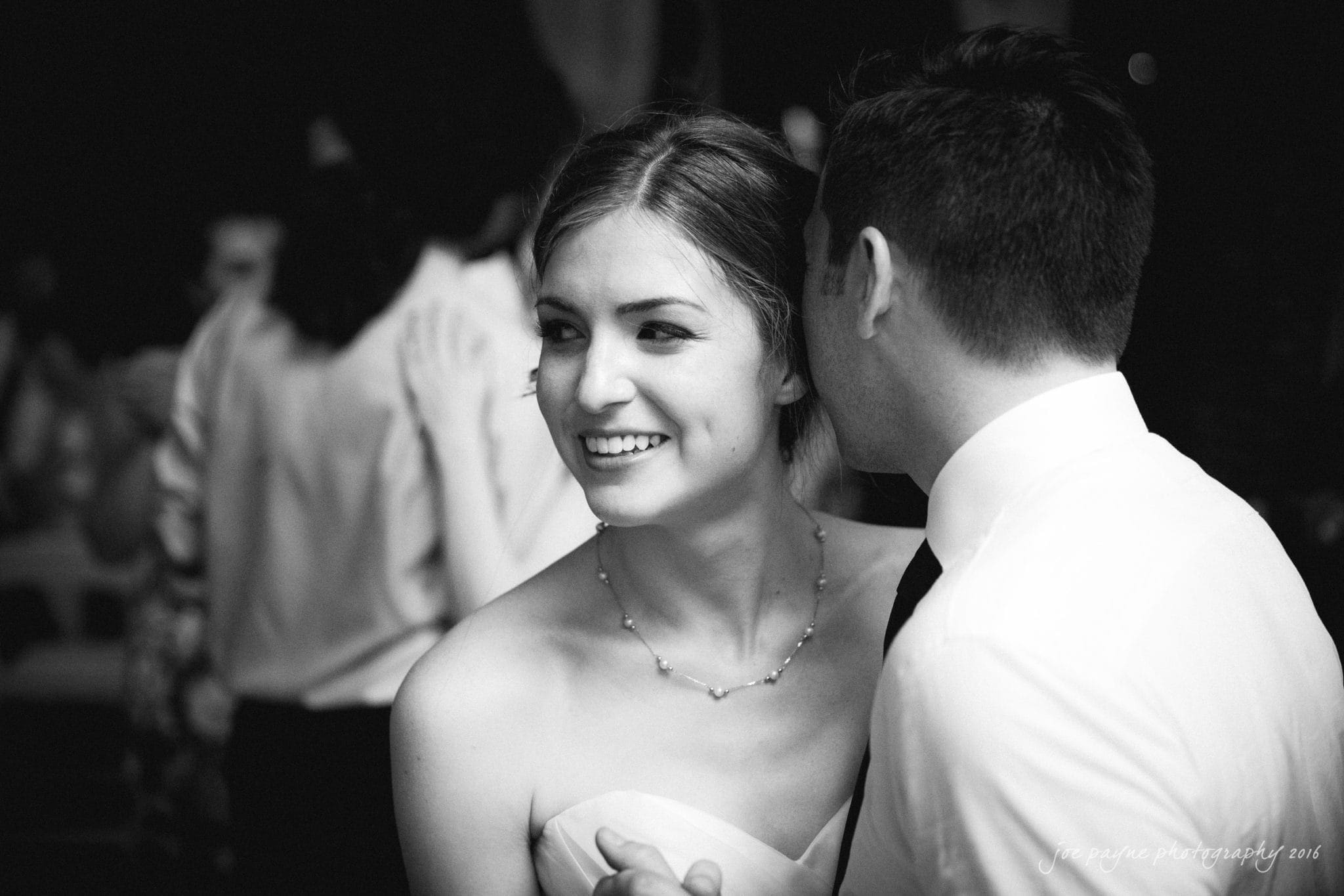 the oaks at salem wedding photography - kathryn & justin A black and white photo of a smiling woman in a strapless dress, wearing a necklace, while a man whispers to her. They appear to be at a formal event with people in the background.