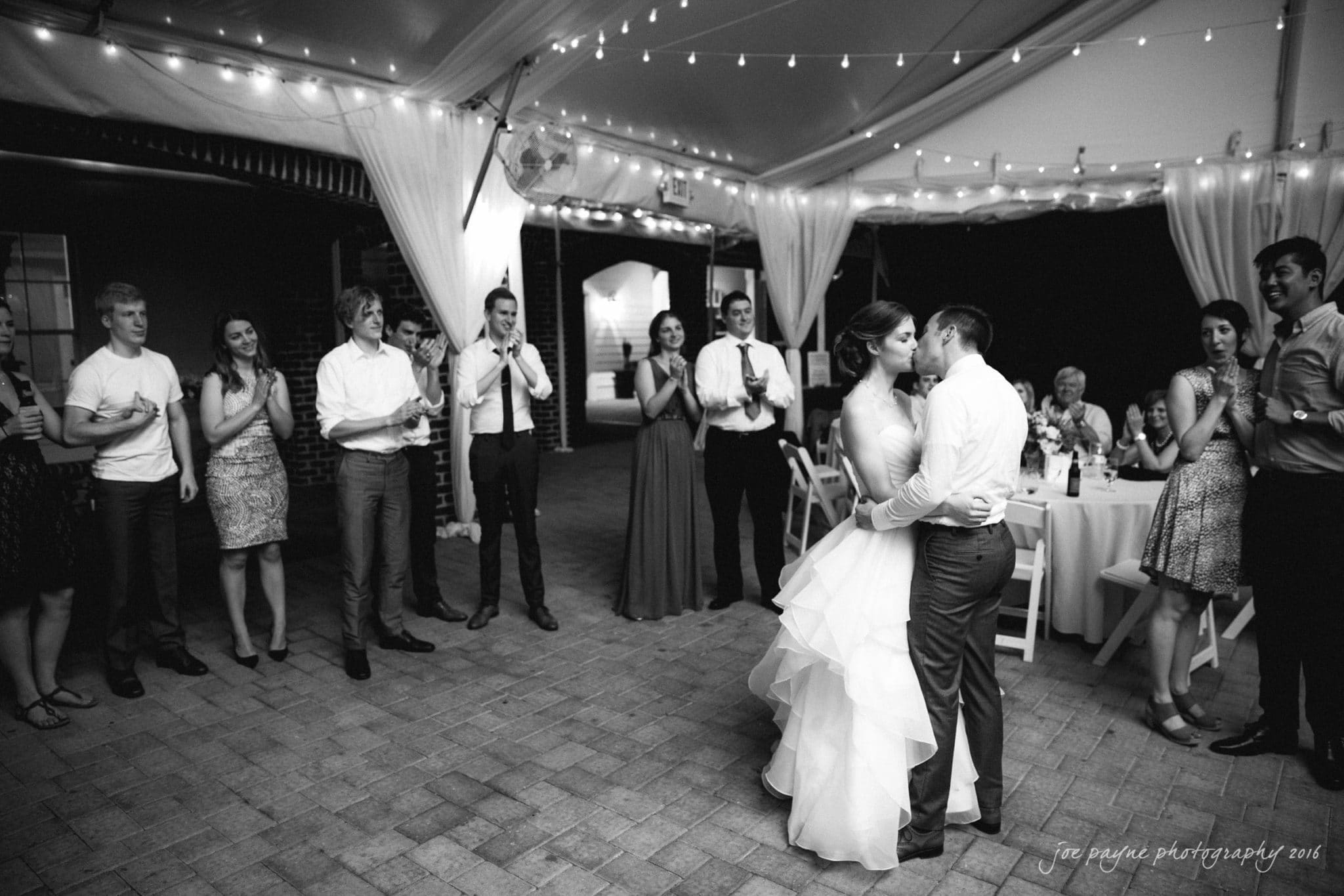 the oaks at salem wedding photography - kathryn & justin A bride and groom share a kiss on the dance floor under a tent with string lights. Guests stand around them clapping and smiling. The setting is elegant and festive.