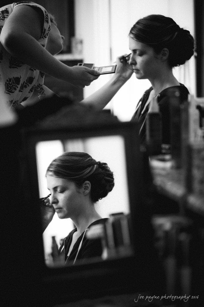 the oaks at salem wedding photography - kathryn & justin A black and white image depicts a woman with an elegant updo having makeup applied. Another reflection in a mirror shows her serene expression. The setting includes makeup tools and a person applying makeup. The atmosphere is calm and intimate.