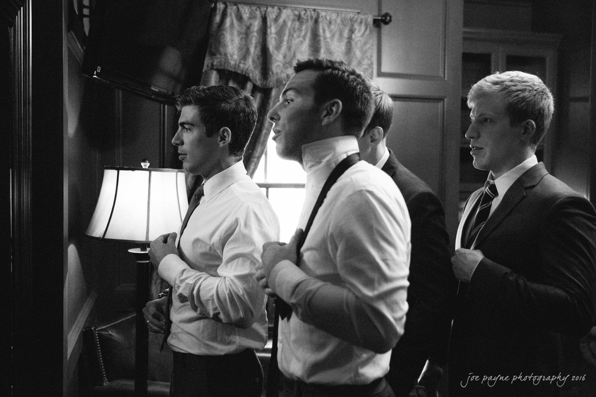 the oaks at salem wedding photography - kathryn & justin A black and white photo of four men in suits adjusting their ties in front of a mirror. They are in a dimly lit room, with a lamp and curtains providing a soft background.