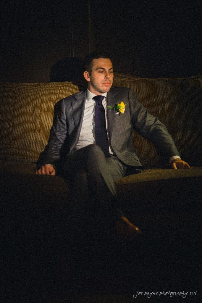 the oaks at salem wedding photography - kathryn & justin A man in a suit sits on a couch, legs crossed, in low lighting. He wears a white shirt, dark tie, and has a yellow boutonniere. He has short hair and a thoughtful expression, and the background is dimly lit, giving a dramatic effect.