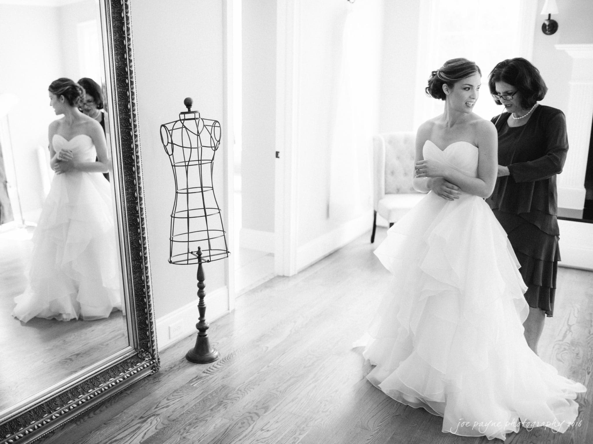 the oaks at salem wedding photography - kathryn & justin A bride in a strapless, ruffled gown is being helped by a woman in a dark dress. They stand in an elegant room with hardwood floors, a large mirror, and a dress form. The atmosphere is serene and joyful.