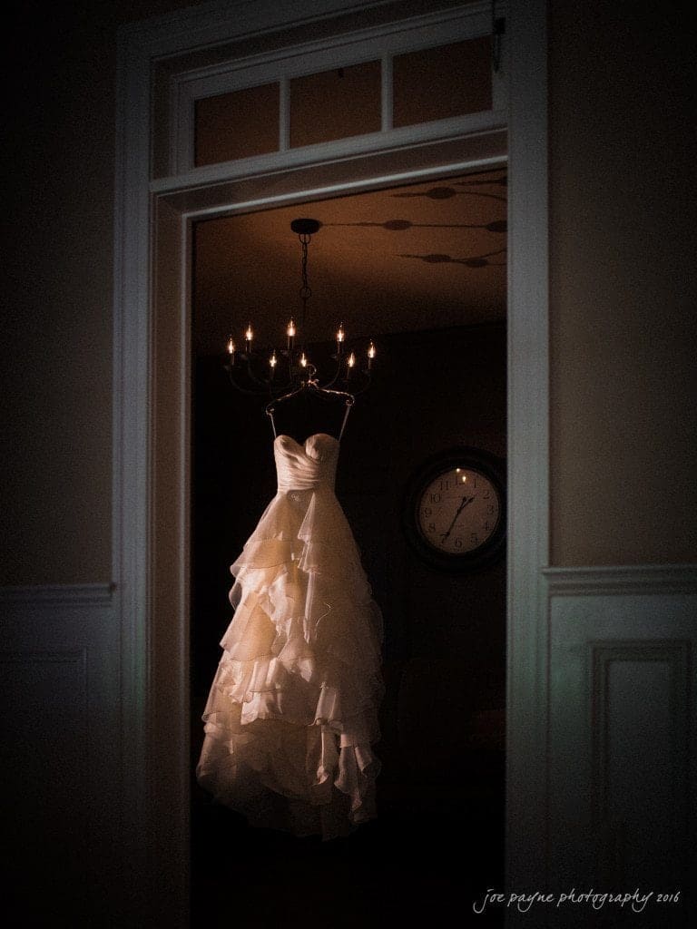 the oaks at salem wedding photography - kathryn & justin A white, strapless wedding dress with layered ruffles hangs from a chandelier in a dimly lit room. A round clock is visible on the wall in the background.