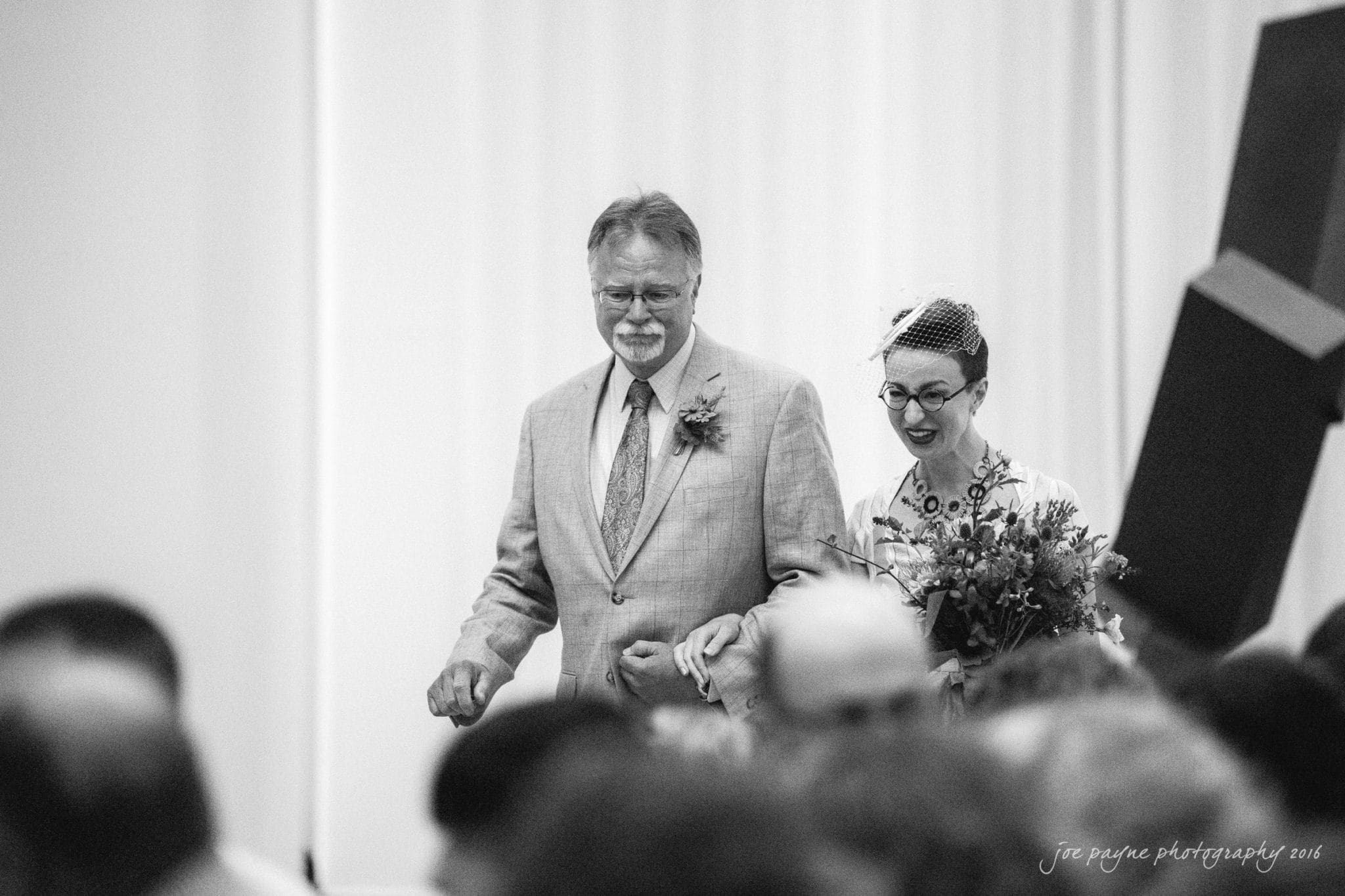 nc museum of art wedding photography - jade & frank A black and white photo of a smiling bride and an older man, possibly her father. The bride holds a bouquet and wears a veil. They walk arm in arm, with people in blurred foreground. The setting appears to be a formal indoor event.