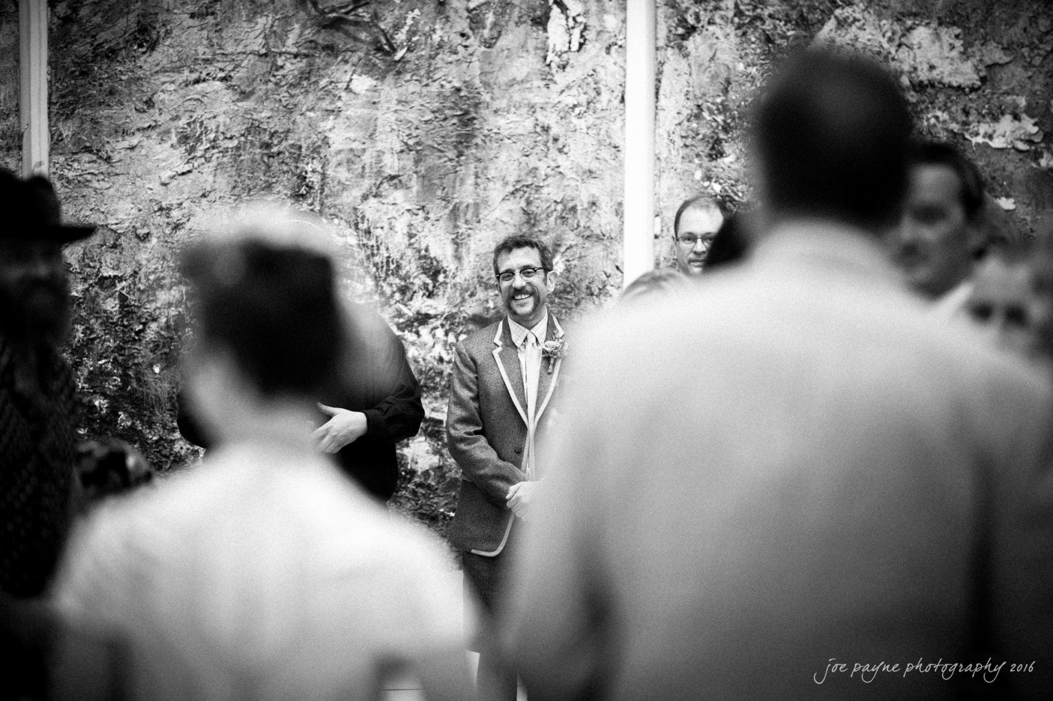 nc museum of art wedding photography - jade & frank A black and white photo of a wedding ceremony. A groom, wearing glasses and a suit, smiles as he stands in front of a textured wall. Blurry figures in the foreground suggest wedding guests or party. The photo has a soft focus and candid feel.