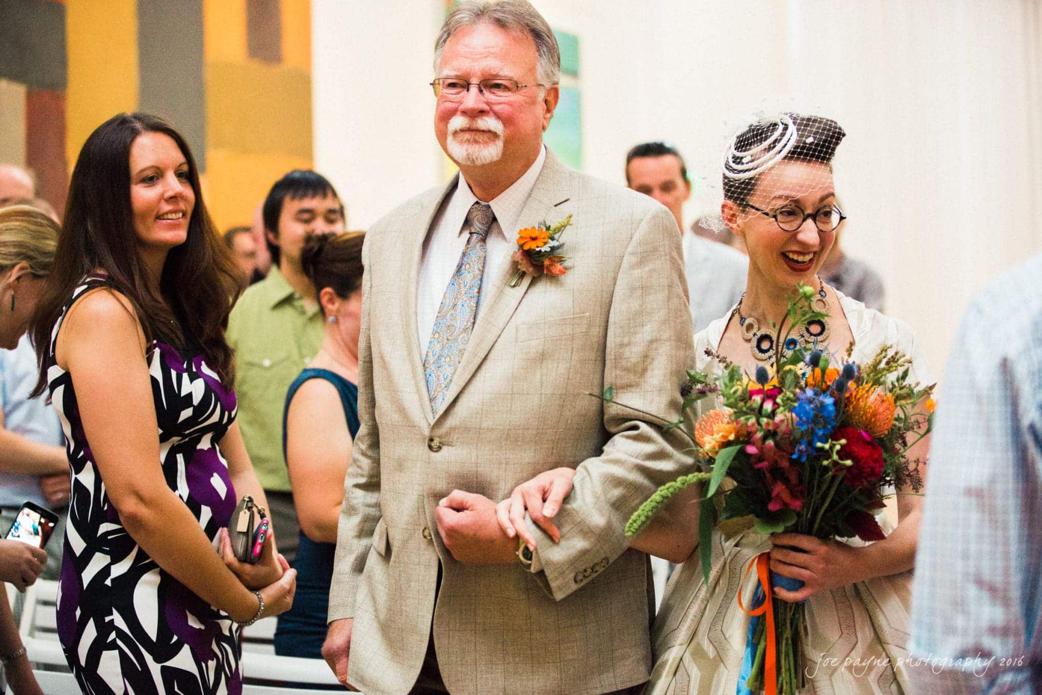 nc museum of art wedding photography - jade & frank A bride in a white dress smiles brightly, holding a colorful bouquet. She stands arm-in-arm with an older man in a beige suit with a floral boutonniere. A woman in a patterned dress stands nearby, surrounded by other guests.