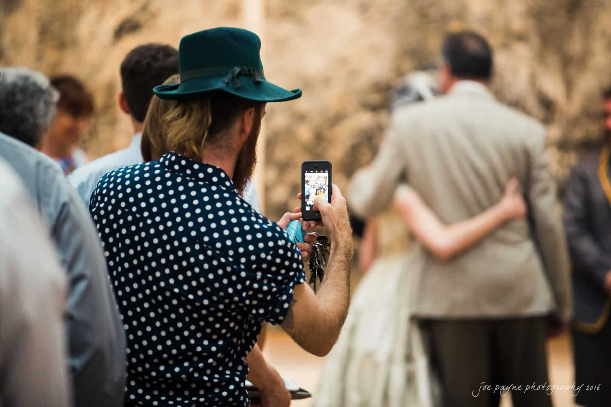 nc museum of art wedding photography - jade & frank A person in a green hat and polka dot shirt takes a photo with a smartphone at an event. A couple poses in the background. The scene appears casual and informal.