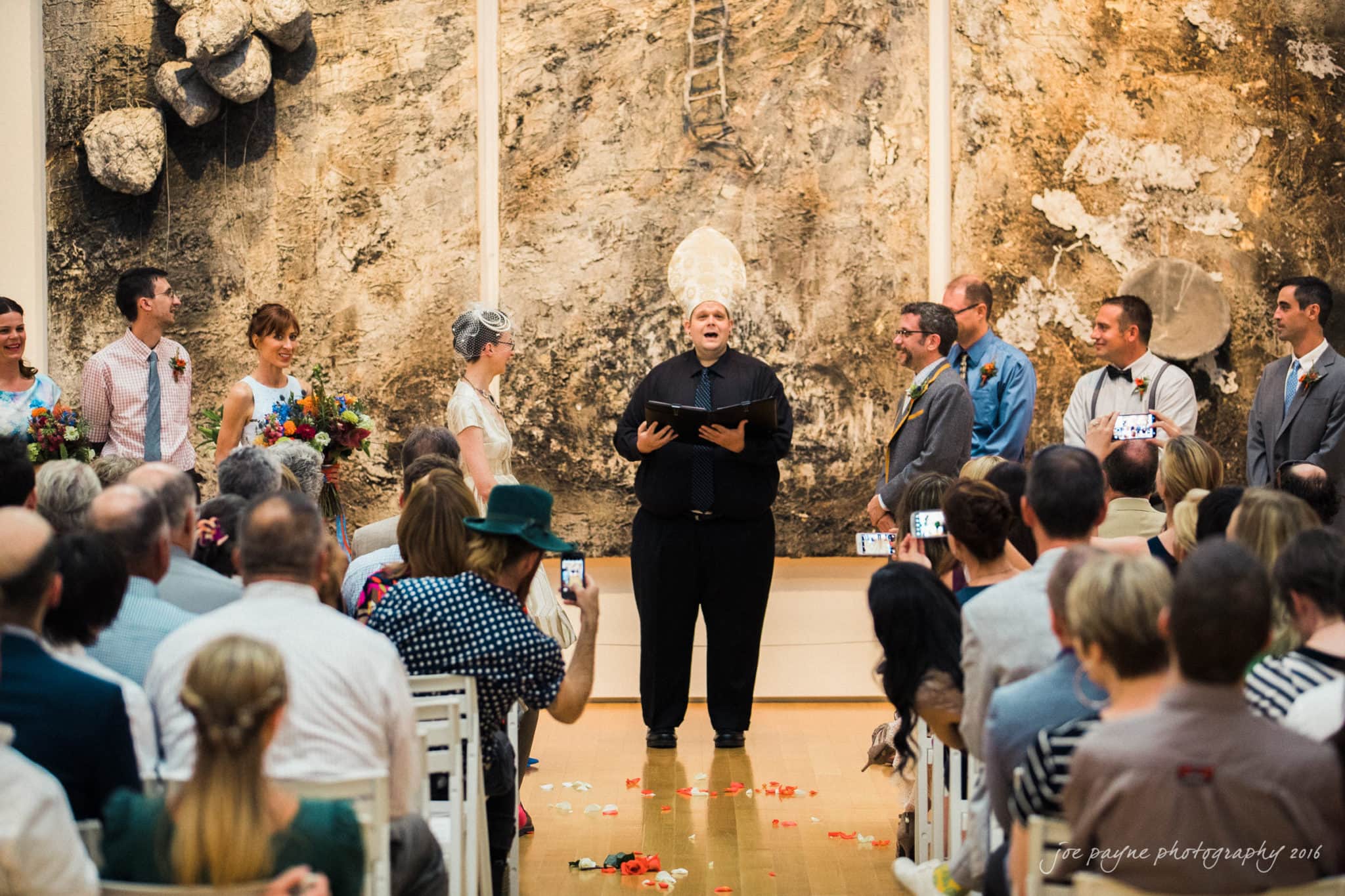 nc museum of art wedding photography - jade & frank A wedding ceremony in an art gallery. The officiant stands in the center, addressing the couple. Bridesmaids and groomsmen are on either side. Guests are seated and taking photos. An abstract artwork is visible in the background.