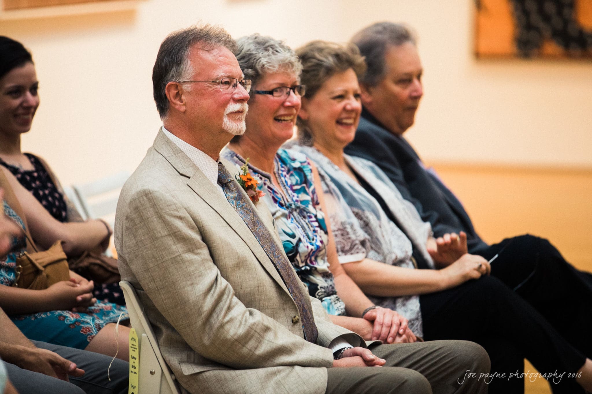 nc museum of art wedding photography - jade & frank A group of five adults sits together on chairs, smiling and laughing. They are dressed in formal and semi-formal attire. The setting appears to be indoors, with an audience in the background.