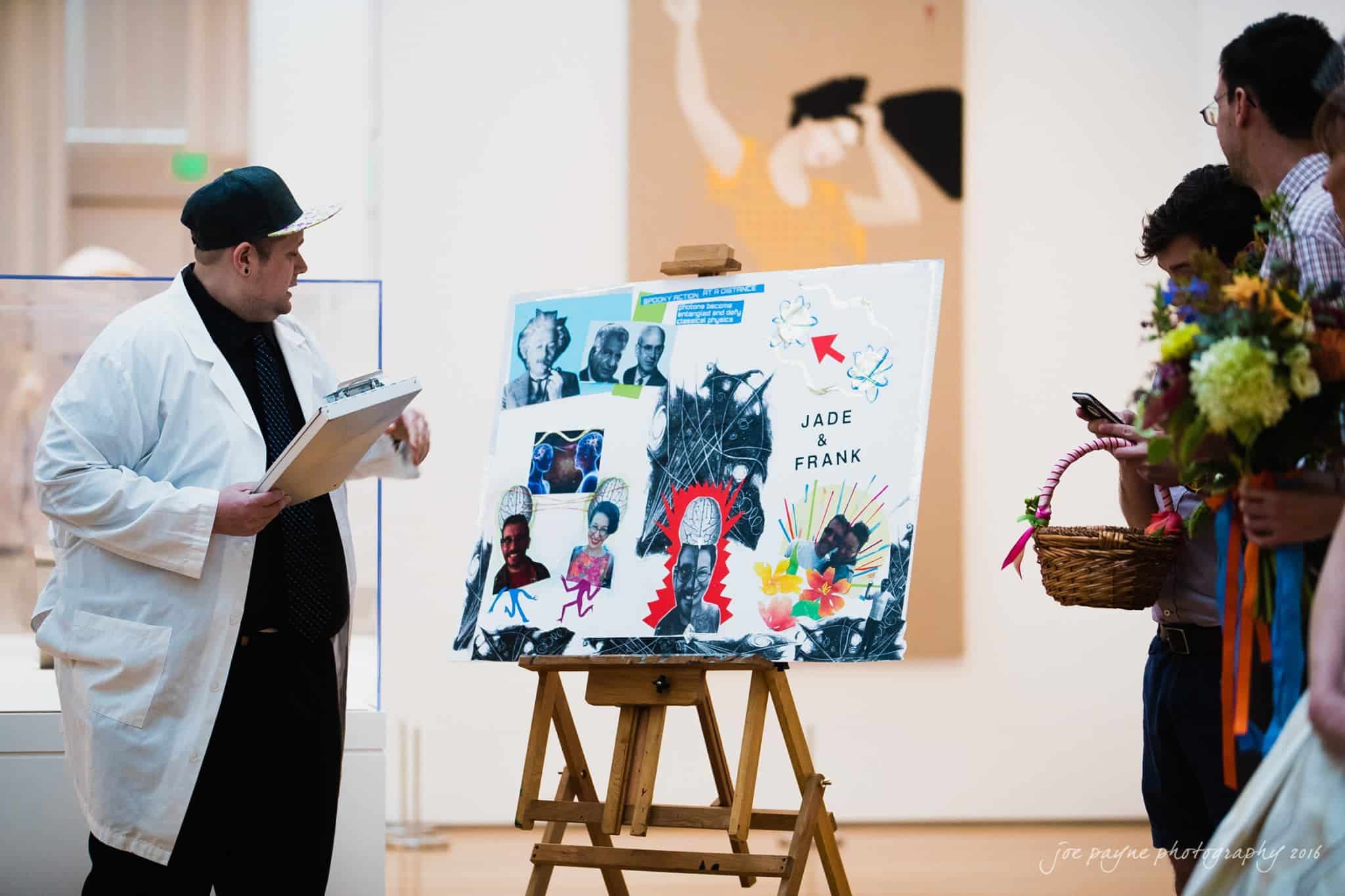nc museum of art wedding photography - jade & frank A man in a white coat and hat is holding a clipboard, standing near an easel displaying a colorful art collage. The collage features various images and text, and several people are nearby, some holding flowers. The setting is a bright gallery space.