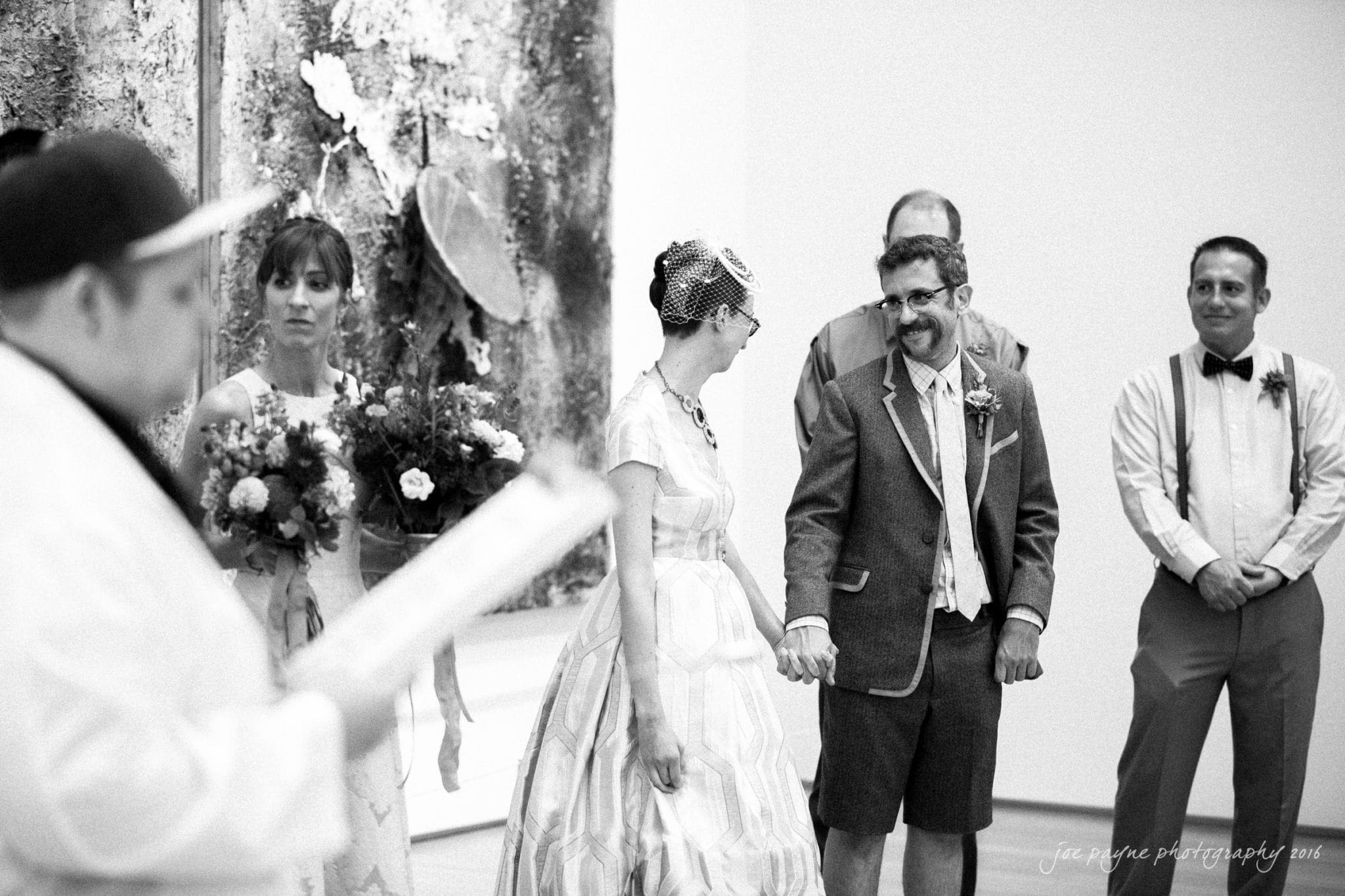 nc museum of art wedding photography - jade & frank A black and white photo of a wedding ceremony. The bride and groom stand holding hands, surrounded by their wedding party. The bride wears a striped dress and veil, the groom wears a suit with shorts. A person in the foreground reads from a document.