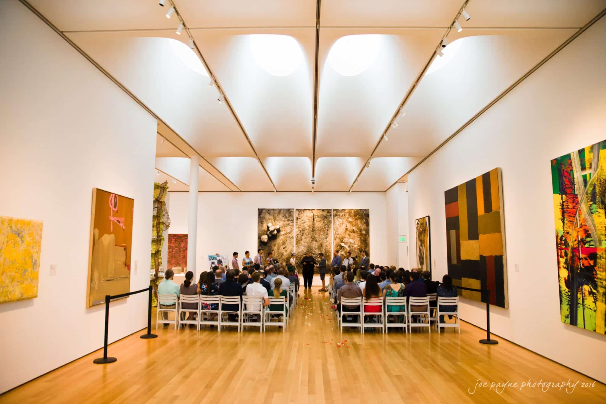nc museum of art wedding photography - jade & frank A spacious art gallery with white walls and a wooden floor displays large, vibrant abstract paintings. A group of people is seated facing a podium, likely for a presentation or event. The ceiling features modern lighting and curved design.