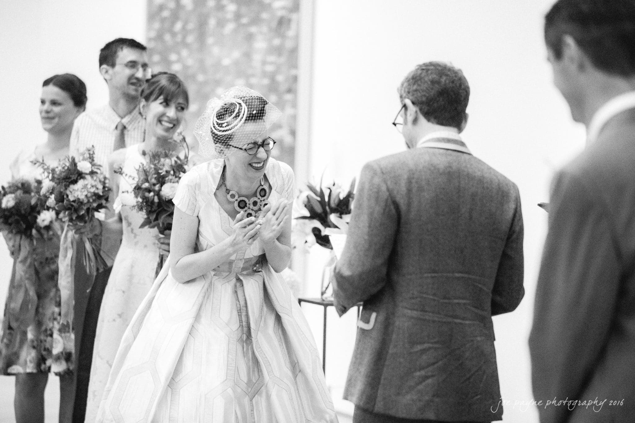 nc museum of art wedding photography - jade & frank A joyful bride in a vintage dress and necklace laughs with a groom in a suit during their wedding ceremony. Bridesmaids and groomsmen stand in the background holding bouquets, all in a bright, elegant setting.