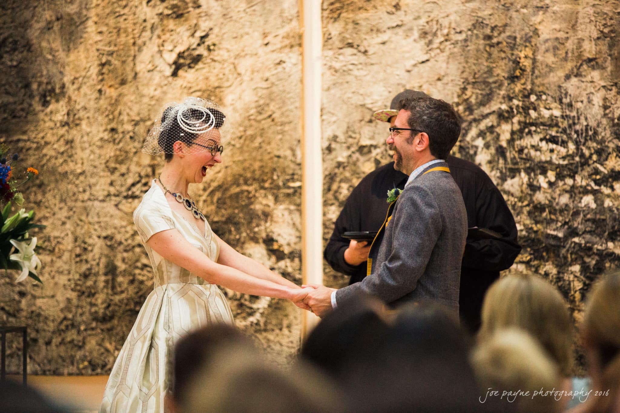 nc museum of art wedding photography - jade & frank A bride in a vintage dress and glasses smiles at the groom, who is wearing a suit with a bow tie. They are holding hands in front of a rustic backdrop during their wedding ceremony. A person stands between them, presumably the officiant.