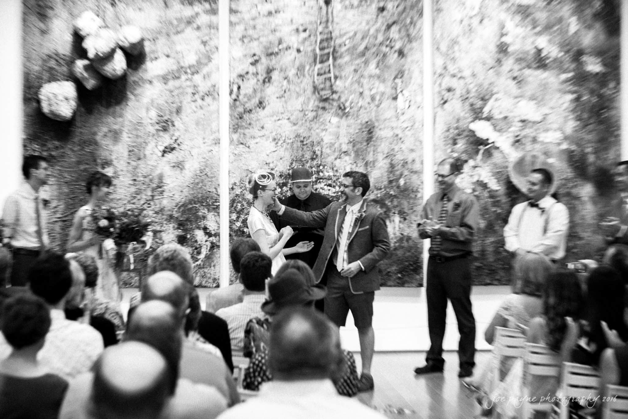nc museum of art wedding photography - jade & frank A black and white photo of a wedding ceremony. The couple stands before an abstract mural. A person between them holds a microphone. Guests are seated, watching the event. The setting appears artistic and intimate.