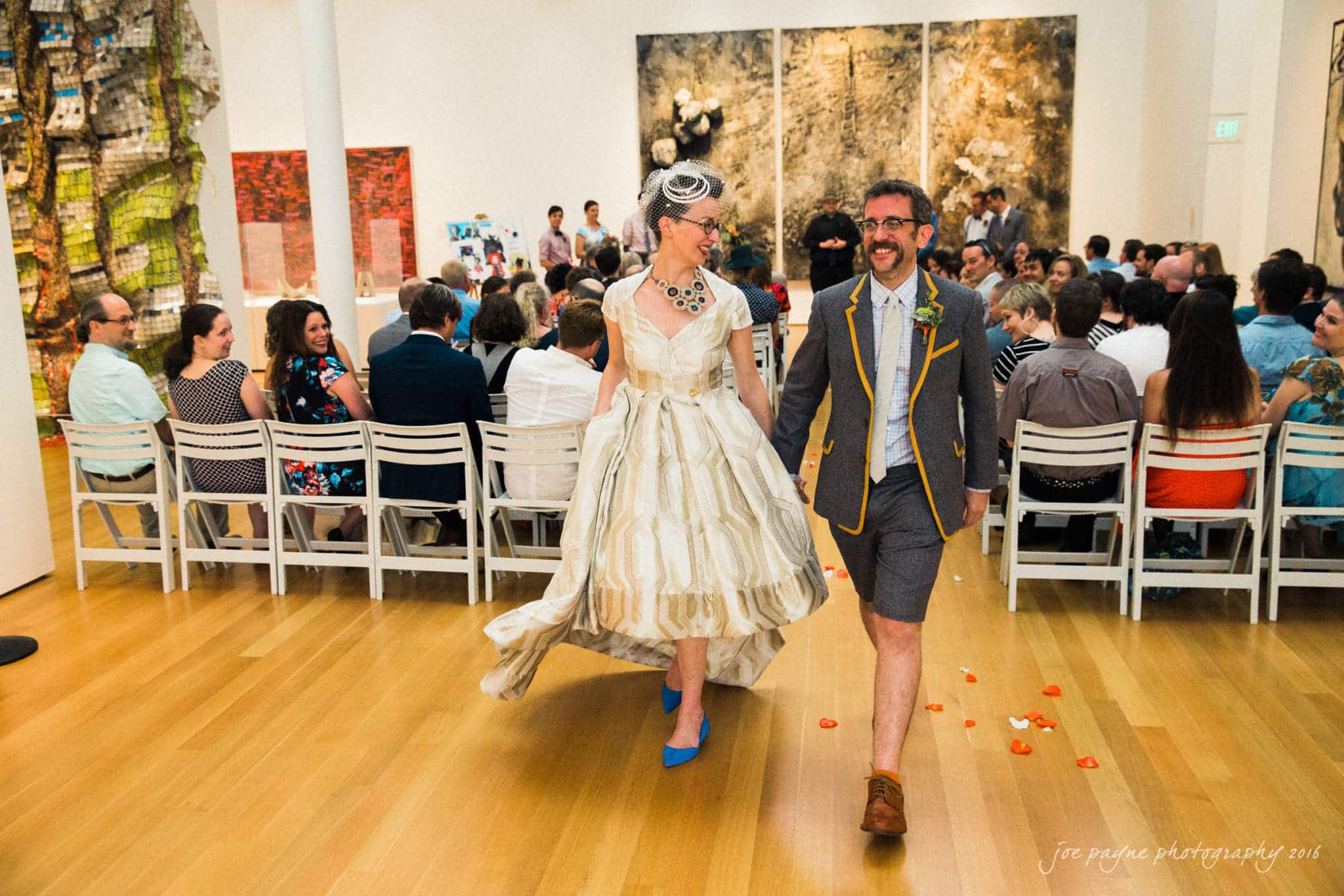 nc museum of art wedding photography - jade & frank A couple joyfully walks down an aisle in an art gallery. The woman is wearing a vintage-style dress and headpiece, and the man is in a gray suit with shorts. Guests are seated and watching. Vibrant art pieces are displayed on the walls.